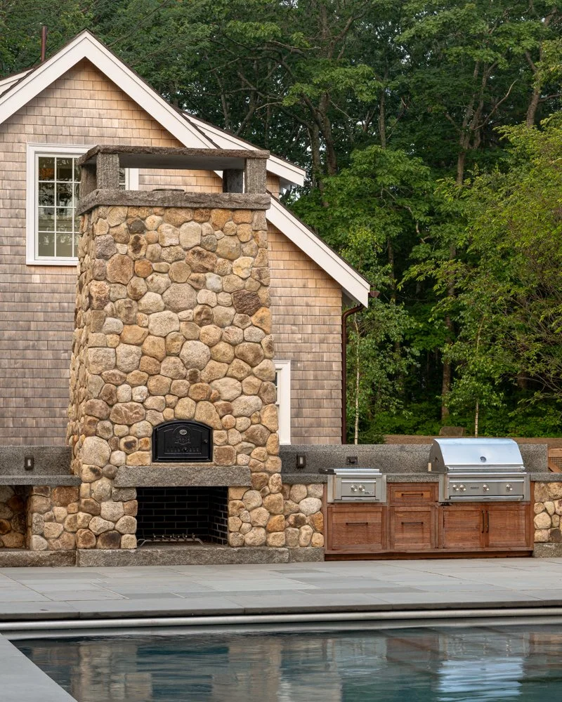 Outdoor kitchen with stone barbecue oven, stainless steel grill, and wood cabinets next to a house with wooden siding surrounded by greenery.
