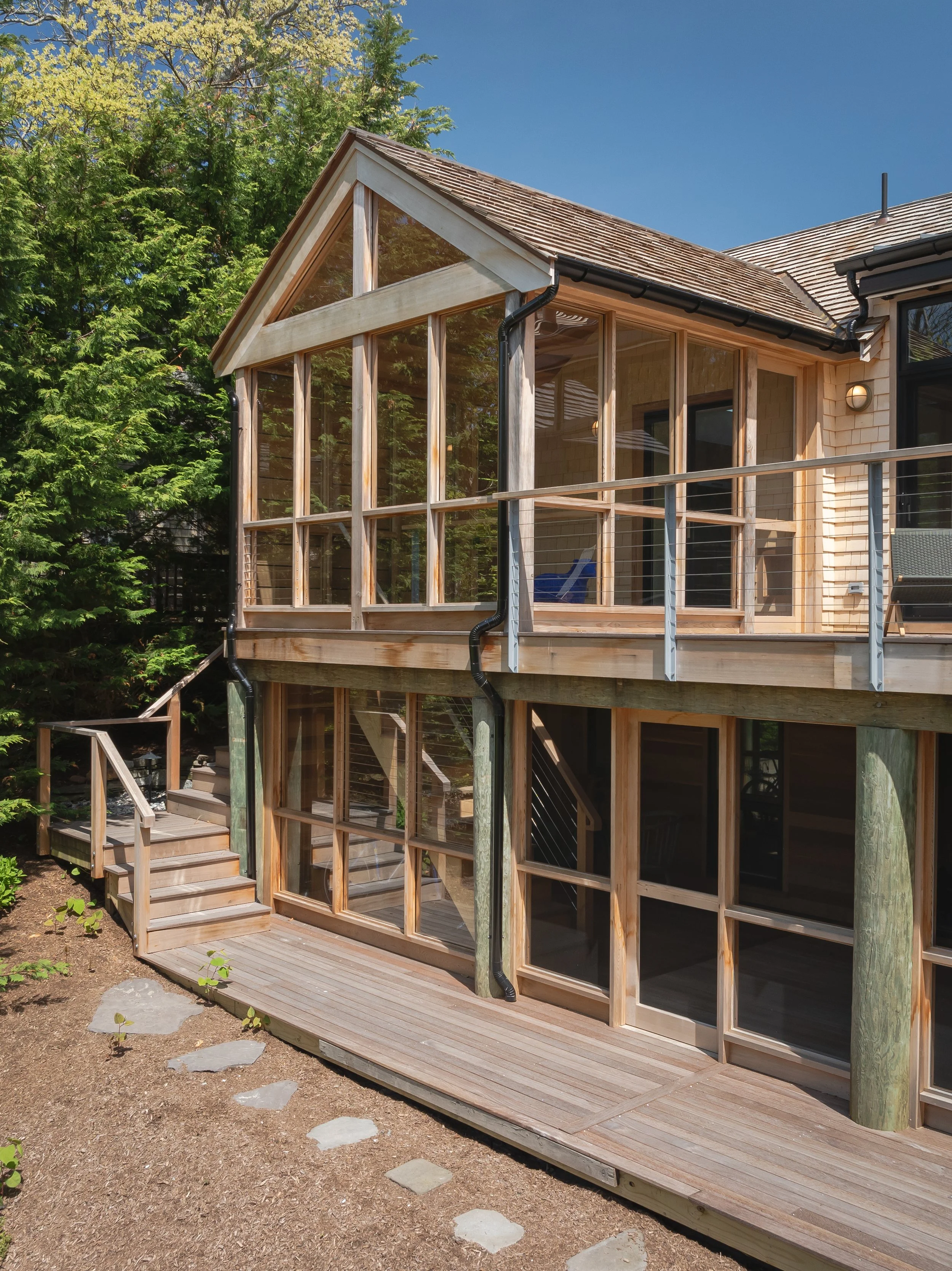 A modern two-story house with extensive wooden framing and large glass windows, surrounded by trees and a clear blue sky.