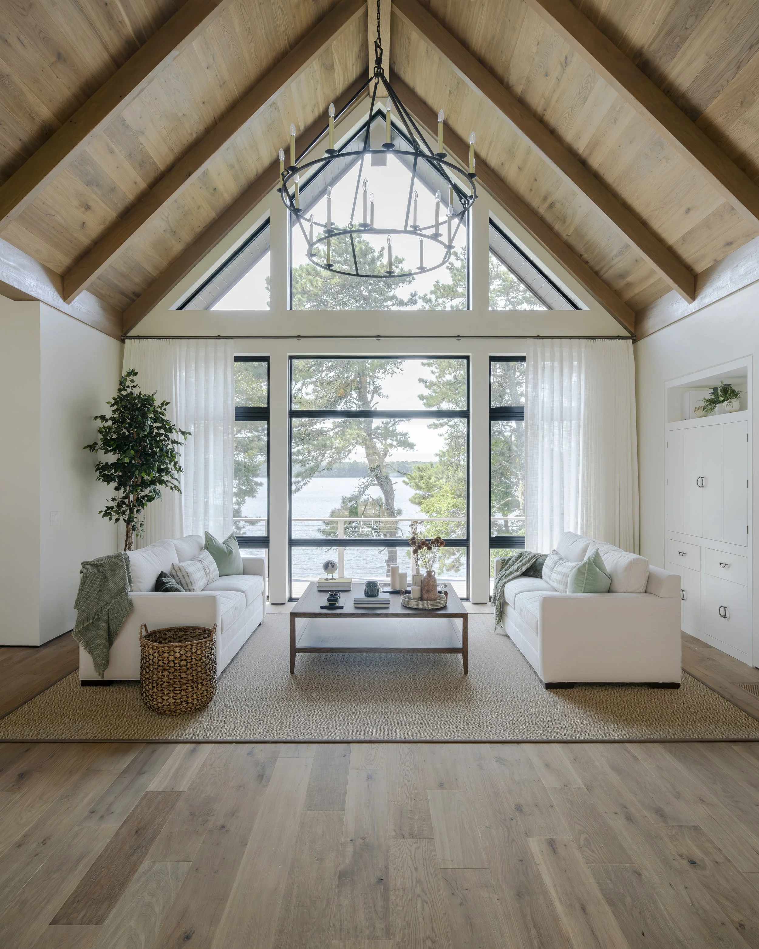 Living room with large windows, white sofas, a wooden coffee table, a potted plant, and a chandelier with candles, overlooking a lakeside view with trees.