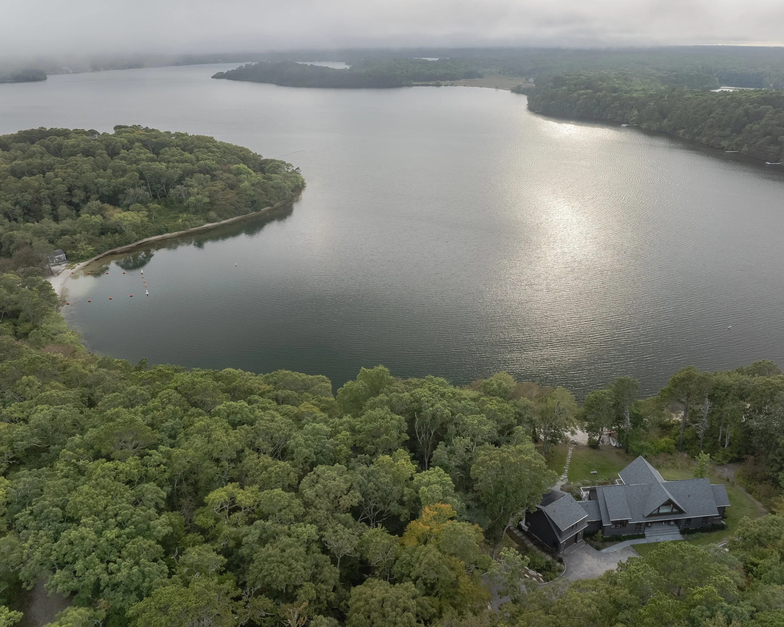 Aerial view of a lake surrounded by dense green forest with a few houses nearby, overcast sky reflecting on the water.