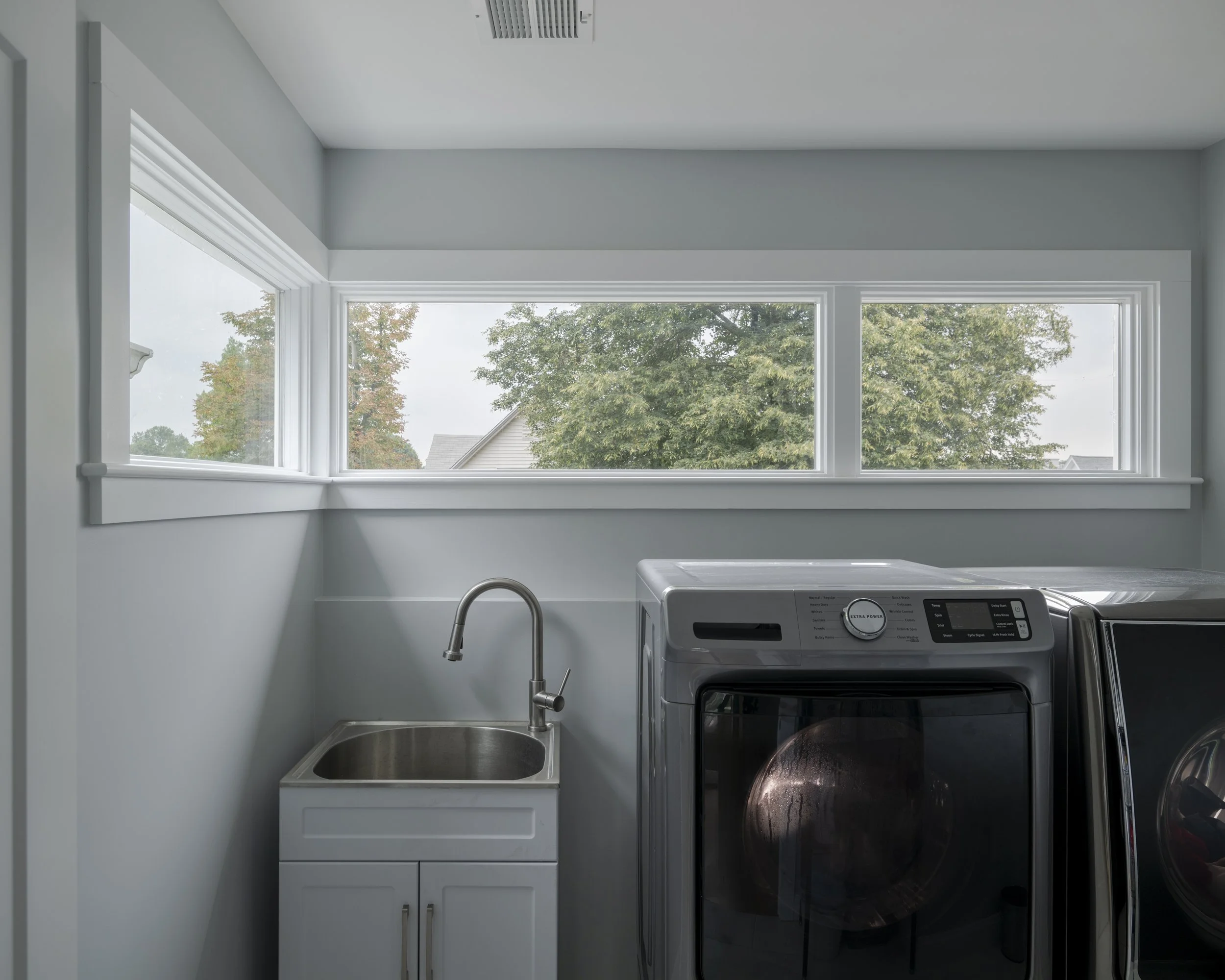 Laundry room with a laundry machine, a sink on a small cabinet, and large windows showing trees outside.