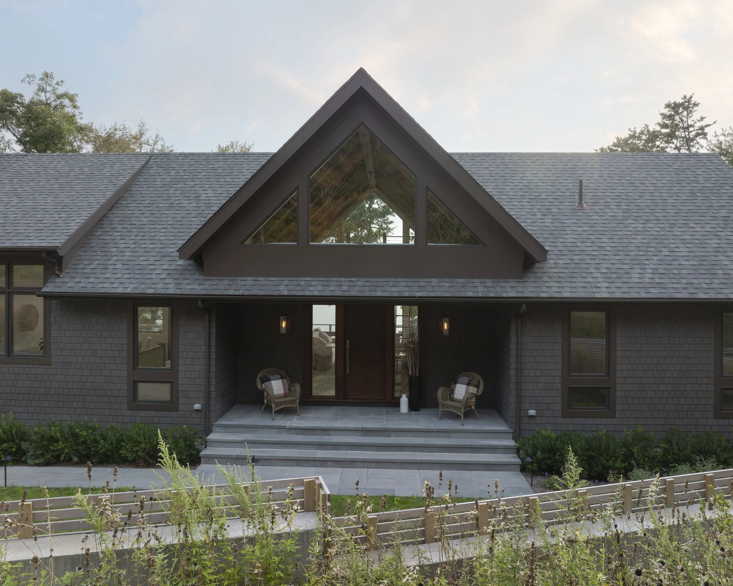 Modern house with dark gray exterior, large triangular window on roof, front porch with two chairs, stairs leading up to entrance, and a landscaped garden in the front.