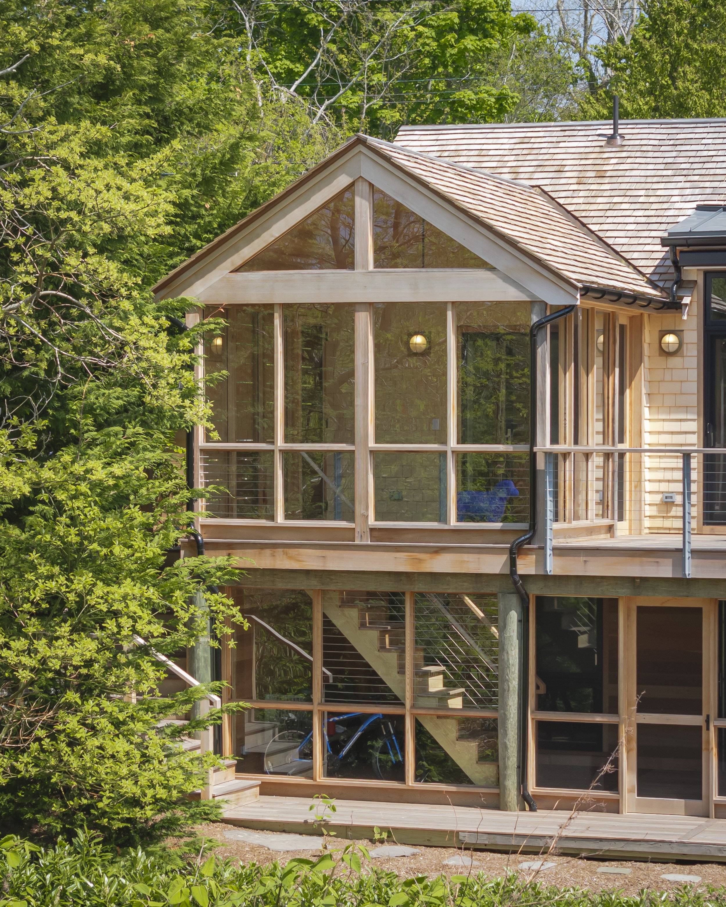 A multi-level house with large glass windows, surrounded by green trees and foliage, featuring a wooden deck and staircase.