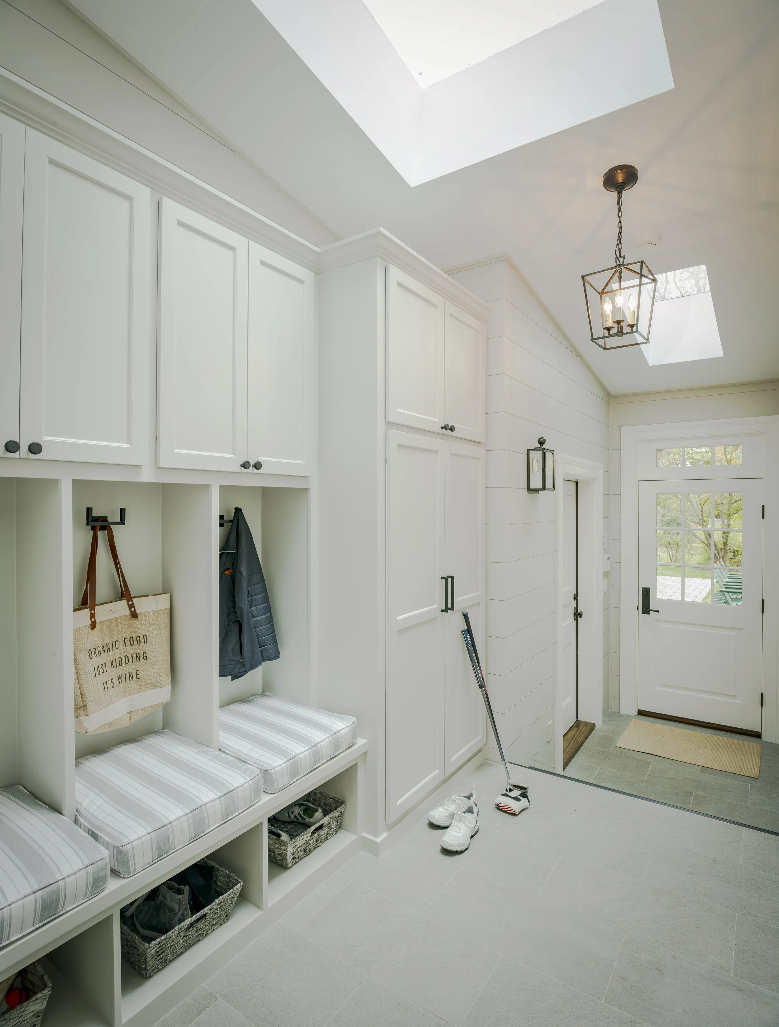 Mudroom with white built-in cubbies, benches, and cabinets, a pair of white shoes and a putter on the floor, front door with glass panes, skylights, and a ceiling light fixture.
