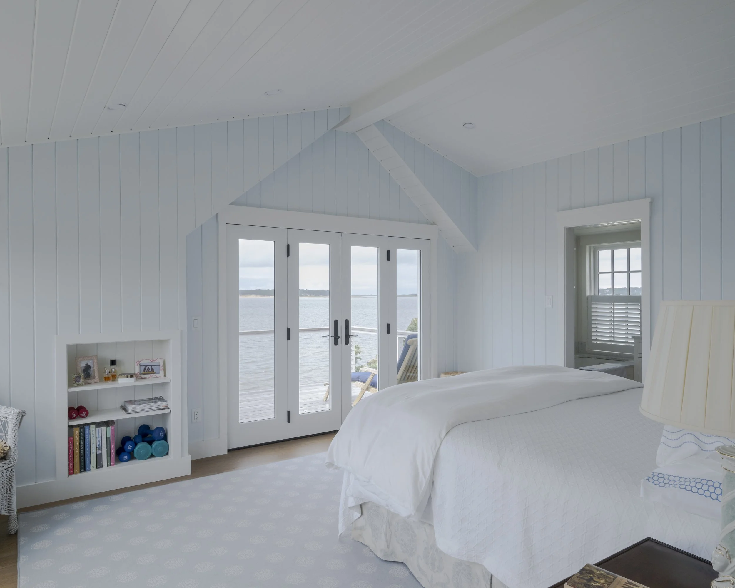 A bright, airy bedroom with white walls, a white bed, and a view of water through glass double doors, with a small bookshelf and a window with white blinds.