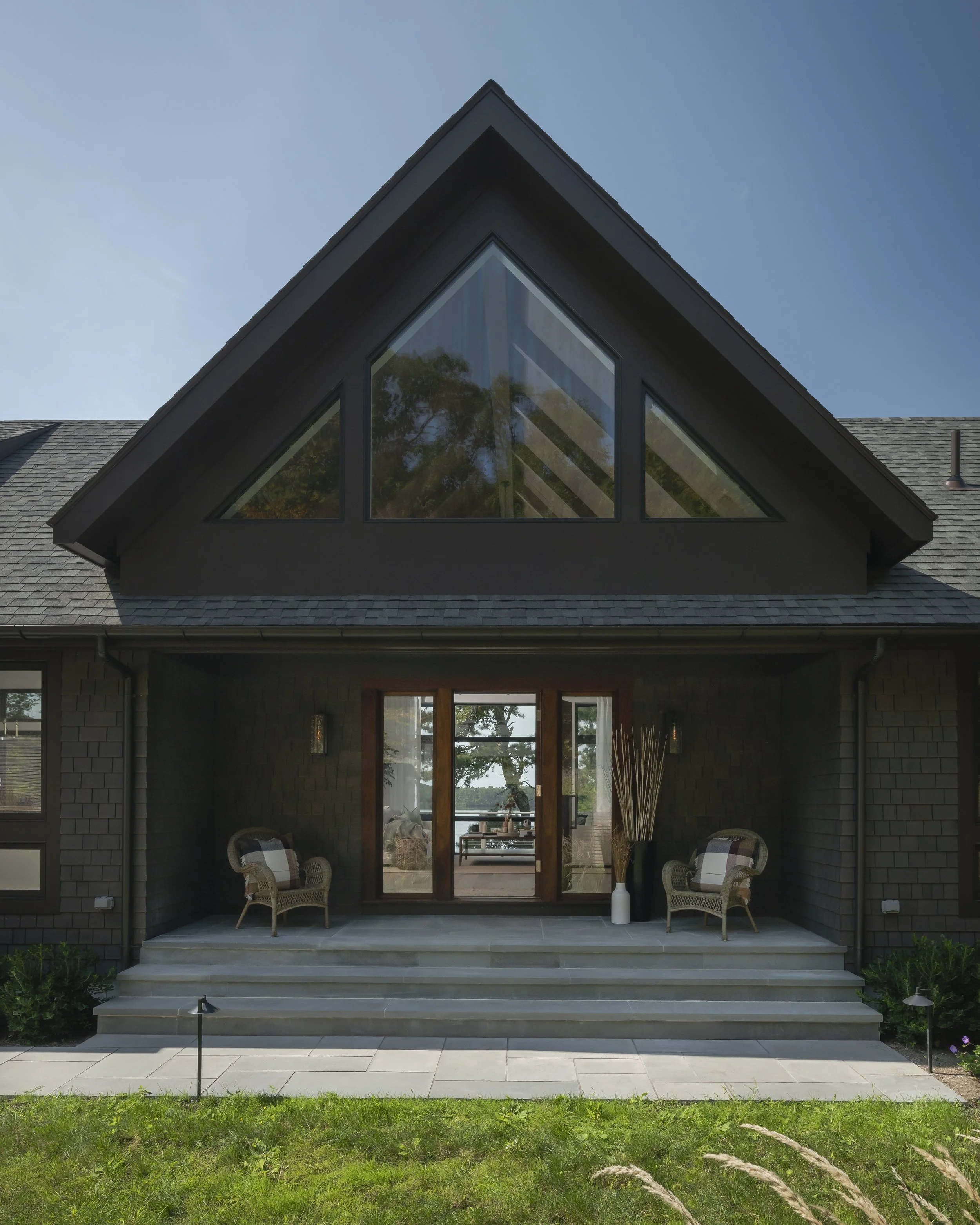 Front view of a modern house with large triangular windows on the upper floor, a wooden sliding door on the porch, and outdoor furniture on the steps.