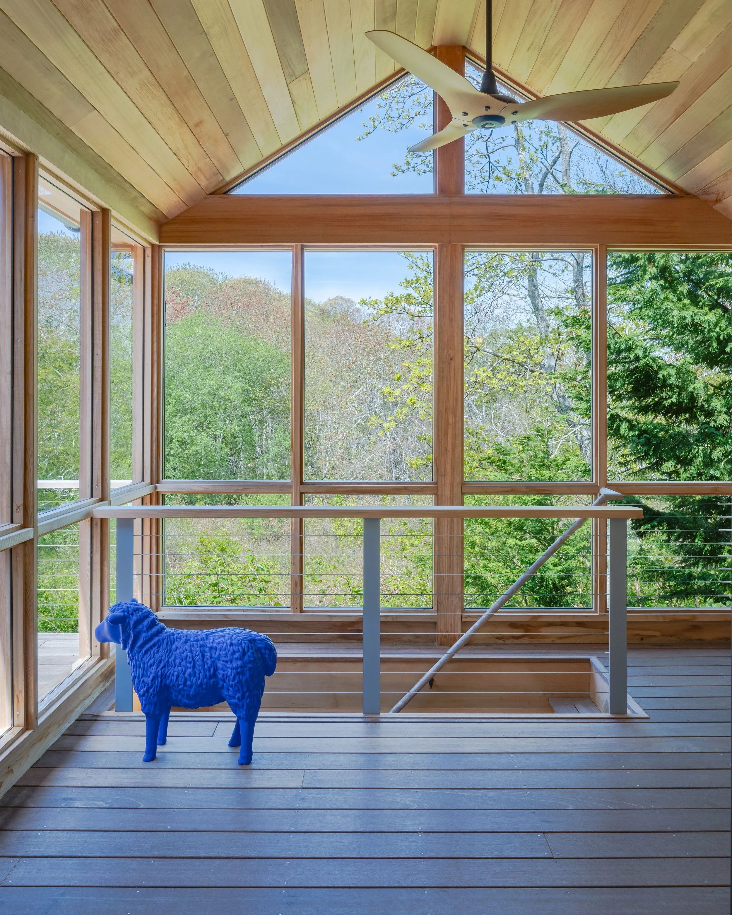 A wooden enclosed balcony with large windows overlooking a green forest, a white ceiling fan, and a blue sheep sculpture.