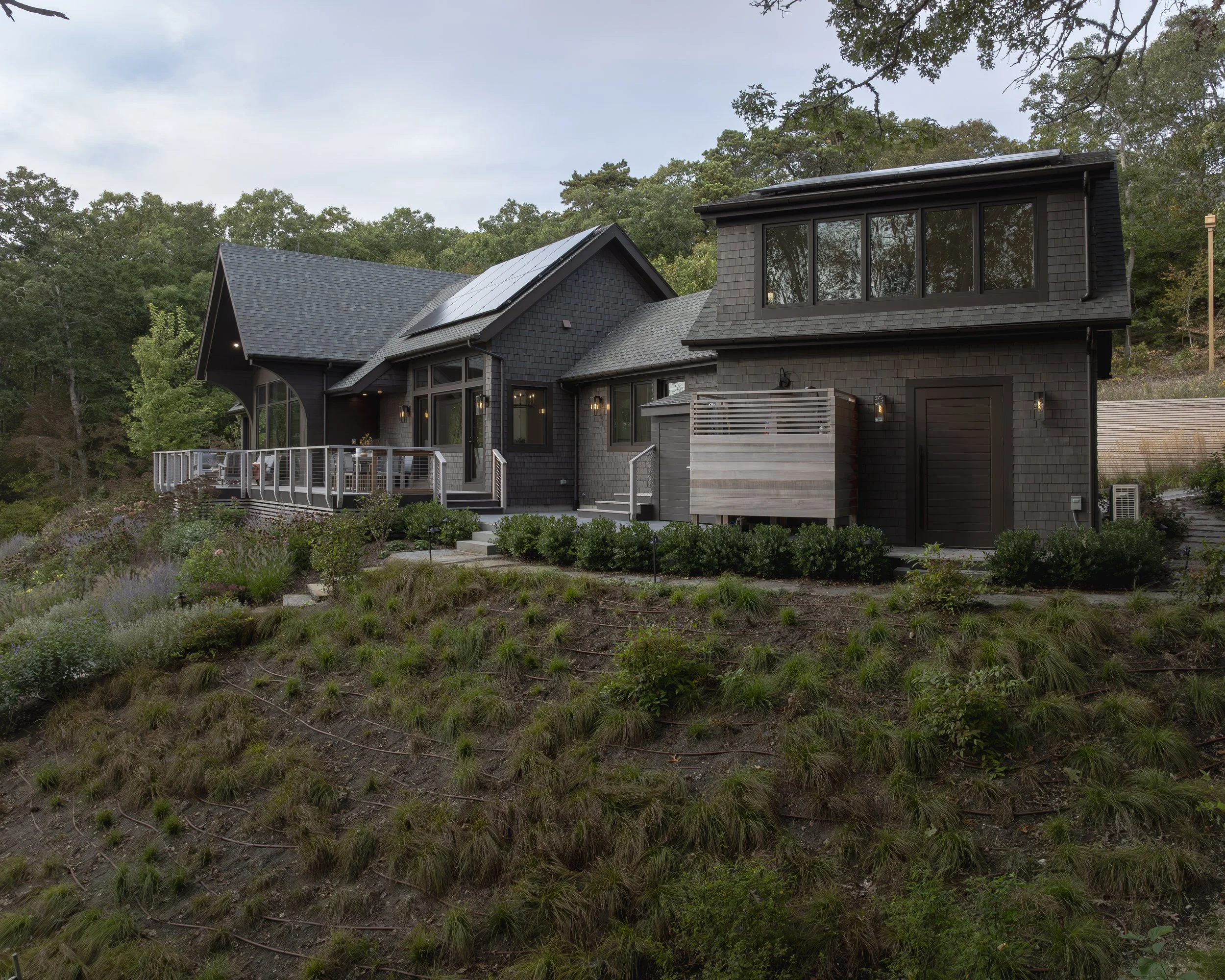 A modern black house with large windows, a deck, and solar panels on the roof, situated on a hillside with a garden and trees in the background.