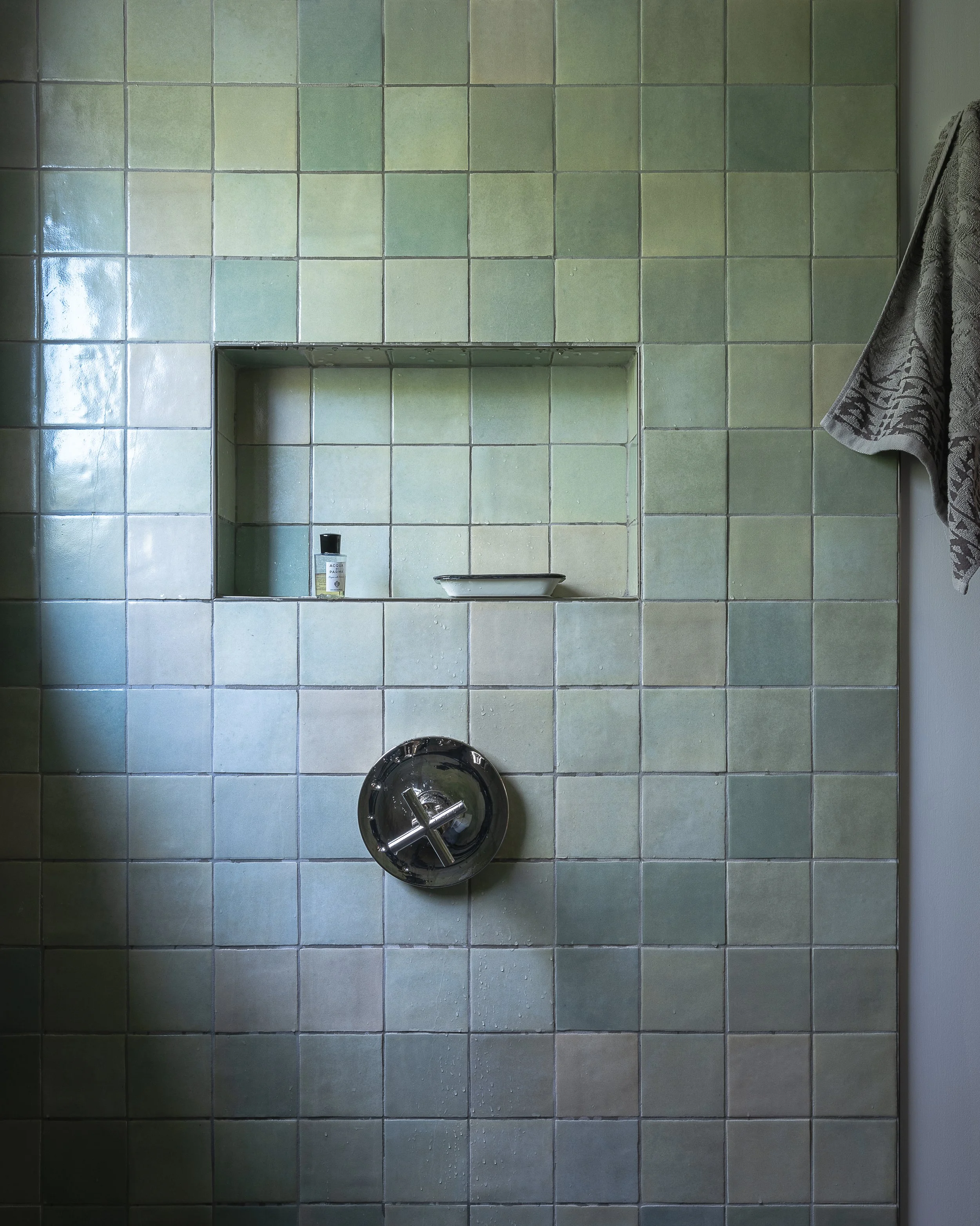A shower wall with green square tiles, a built-in niche with a small bottle and soap dish, and a silver control knob.