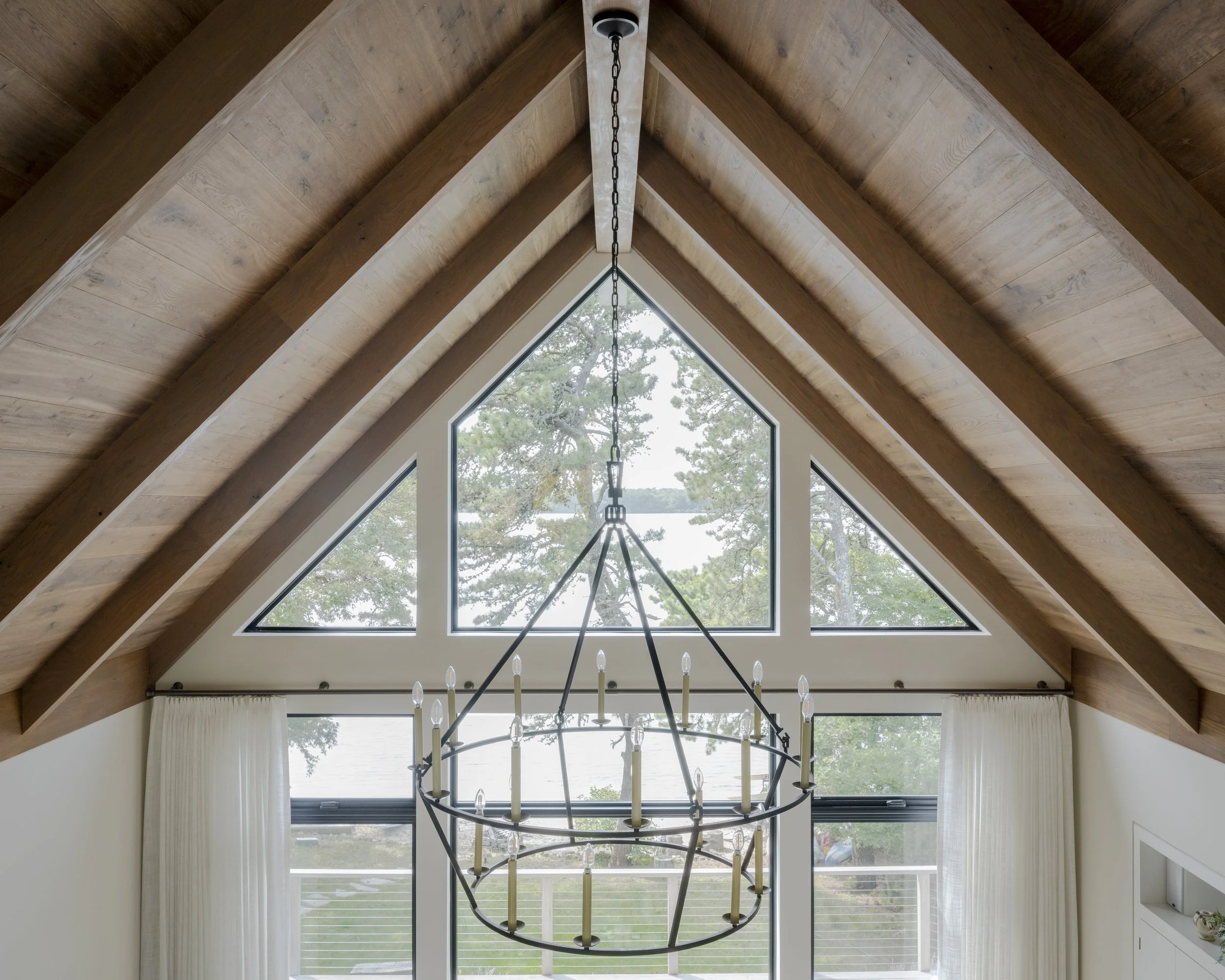 Interior view of a room with high vaulted wooden ceiling, large triangular window, and a black metal chandelier with candles hanging from the ceiling.