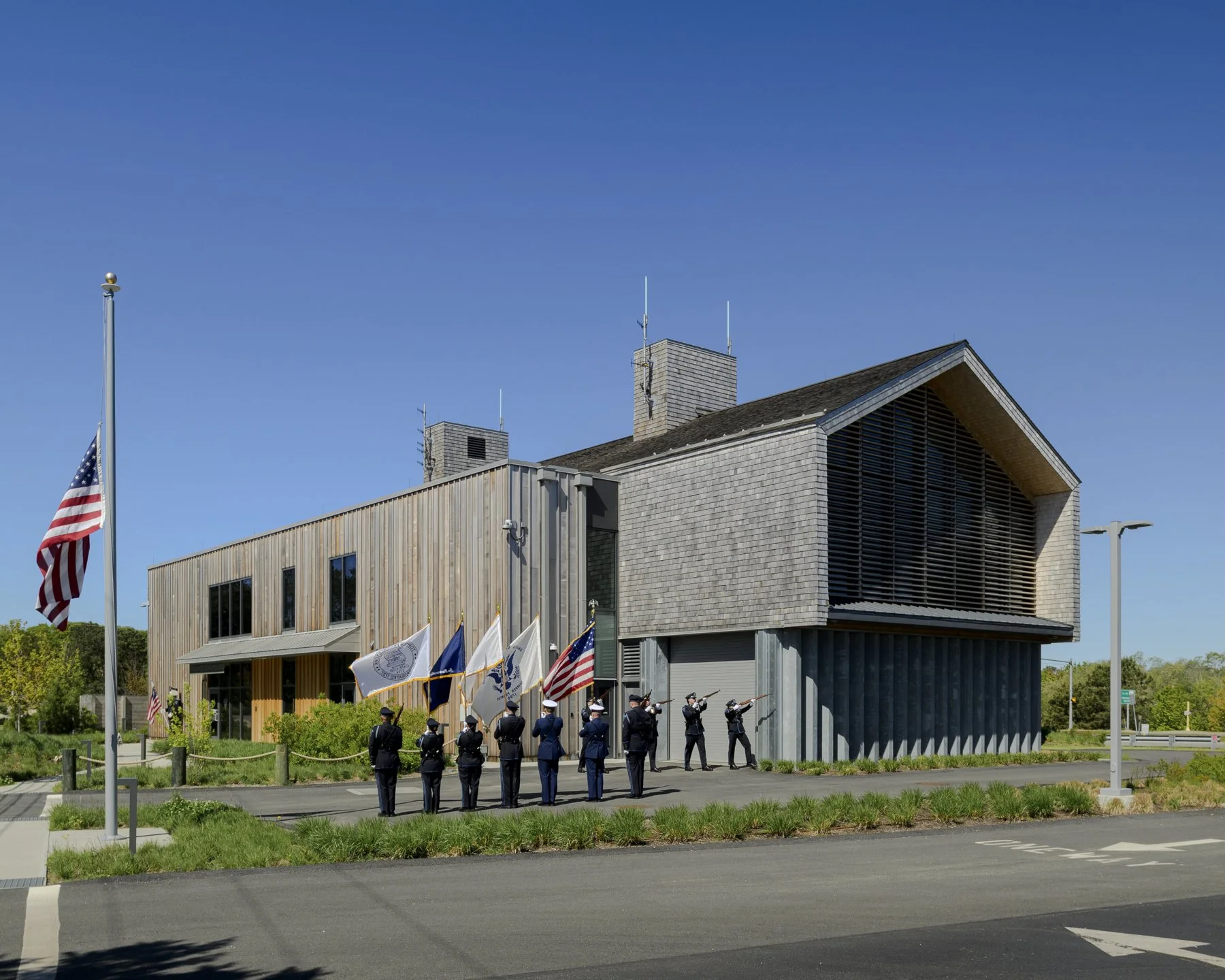 A group of uniformed service members standing and saluting in front of a modern building, with American flags surrounding them, during a flag ceremony on a clear day.