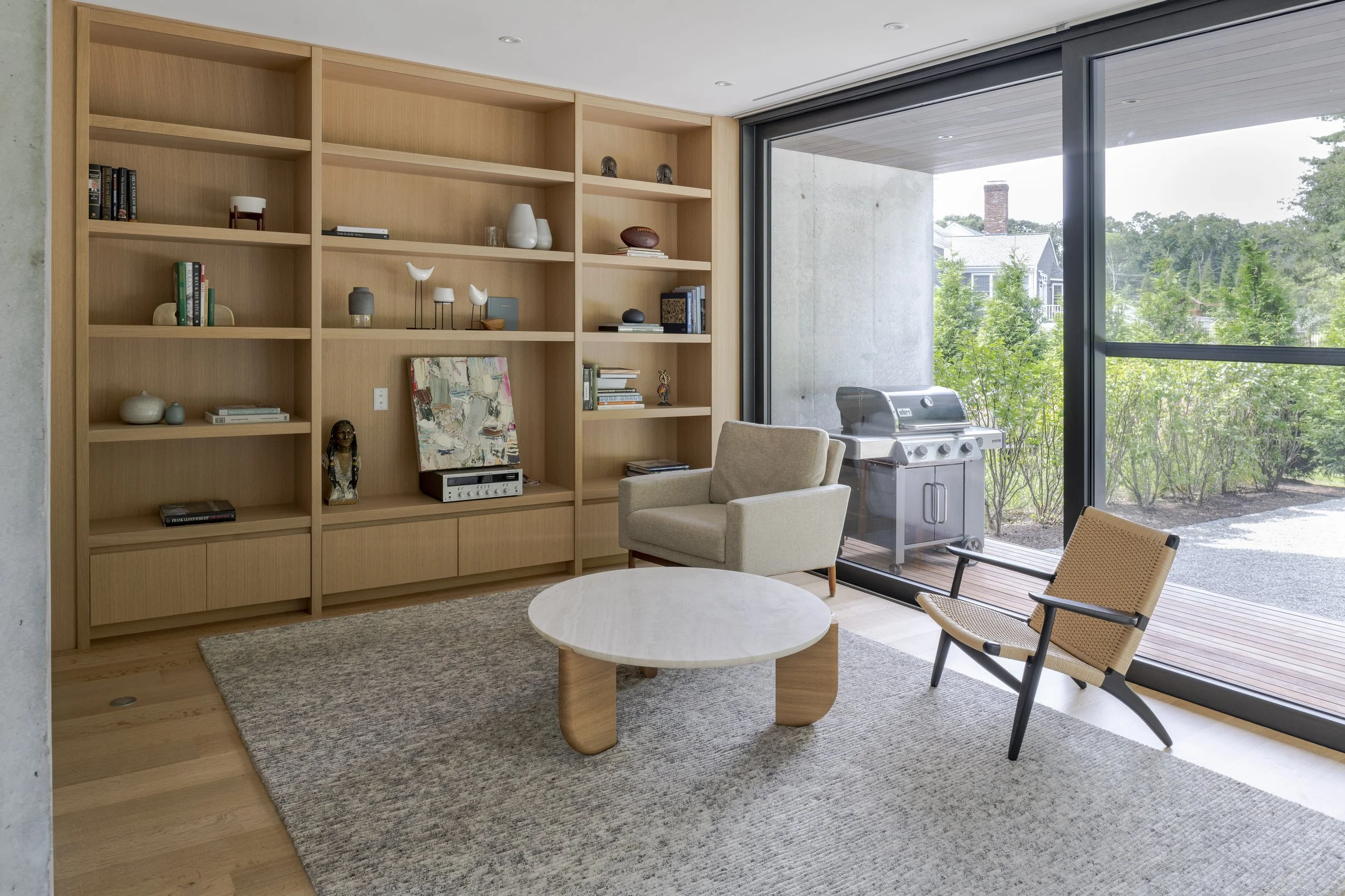 Modern living room with wooden shelf, beige armchair, wooden chair, marble coffee table, gray rug, large sliding glass door with outdoor patio and grill, trees outside.