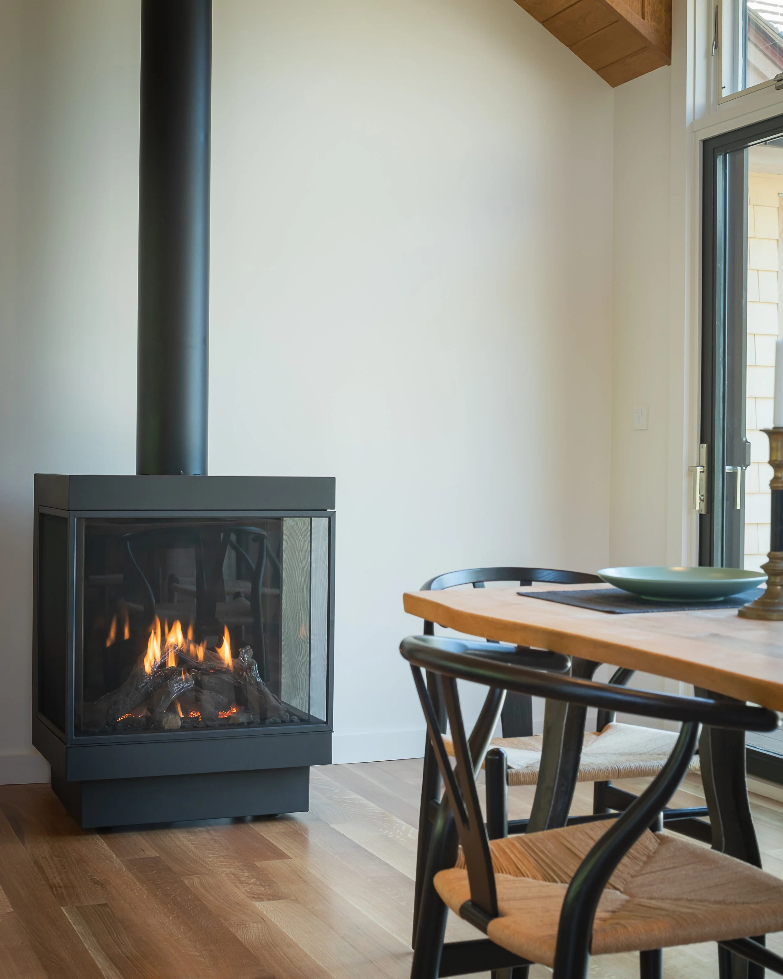 Interior of a room with a modern black fireplace with a glass front and a wood-burning fire inside, alongside a wooden dining table and black chairs, near a sliding glass door and a window.
