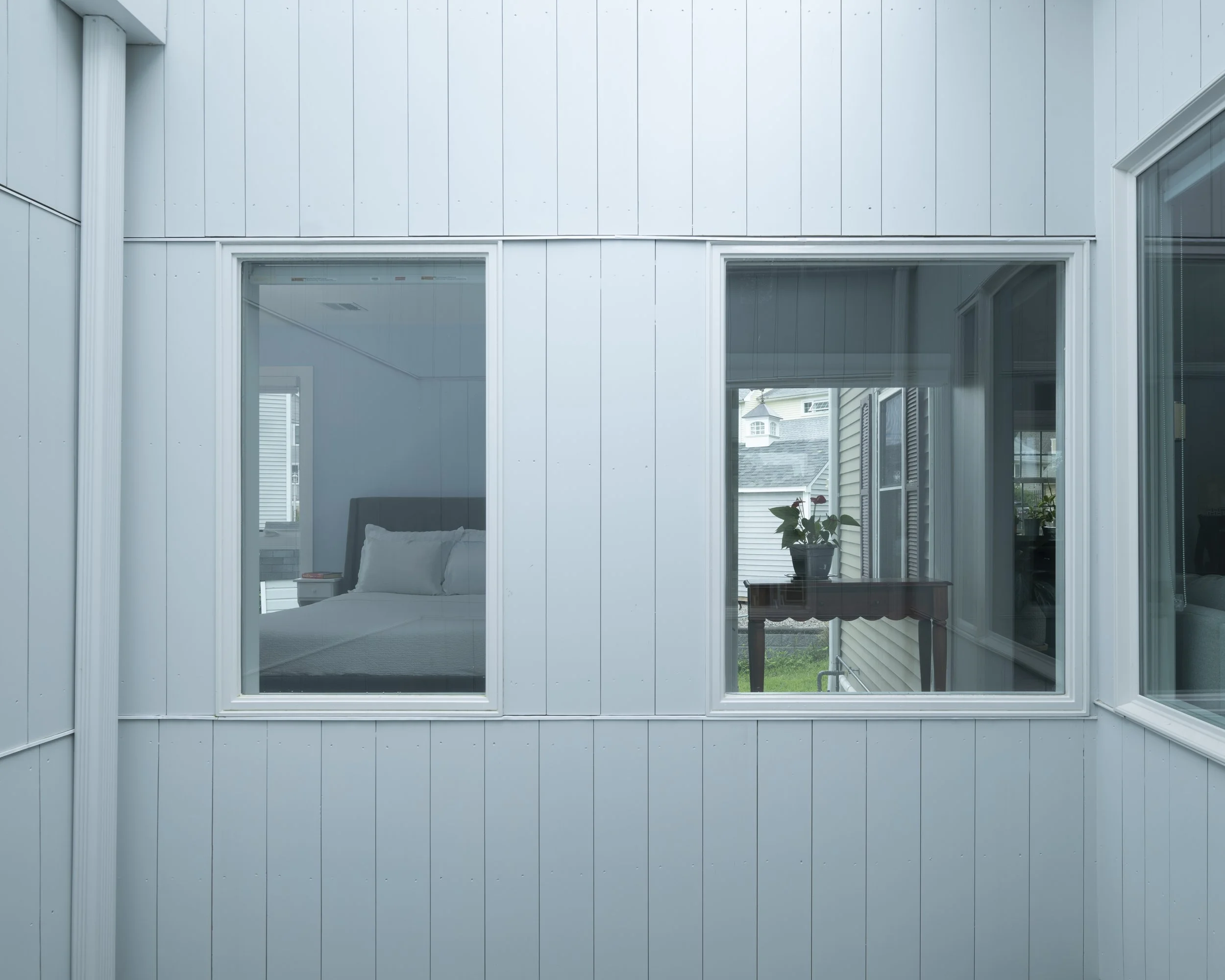 View of a house's exterior wall with three large windows showing the interior and neighboring houses.
