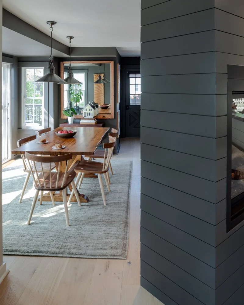 Dining area with a wooden table and six chairs on a light-colored rug, surrounded by windows and a sliding door, with sunlight streaming in. A mirror with a wooden frame, a potted plant, a model house, and a bowl of apples are on the sideboard. Two h