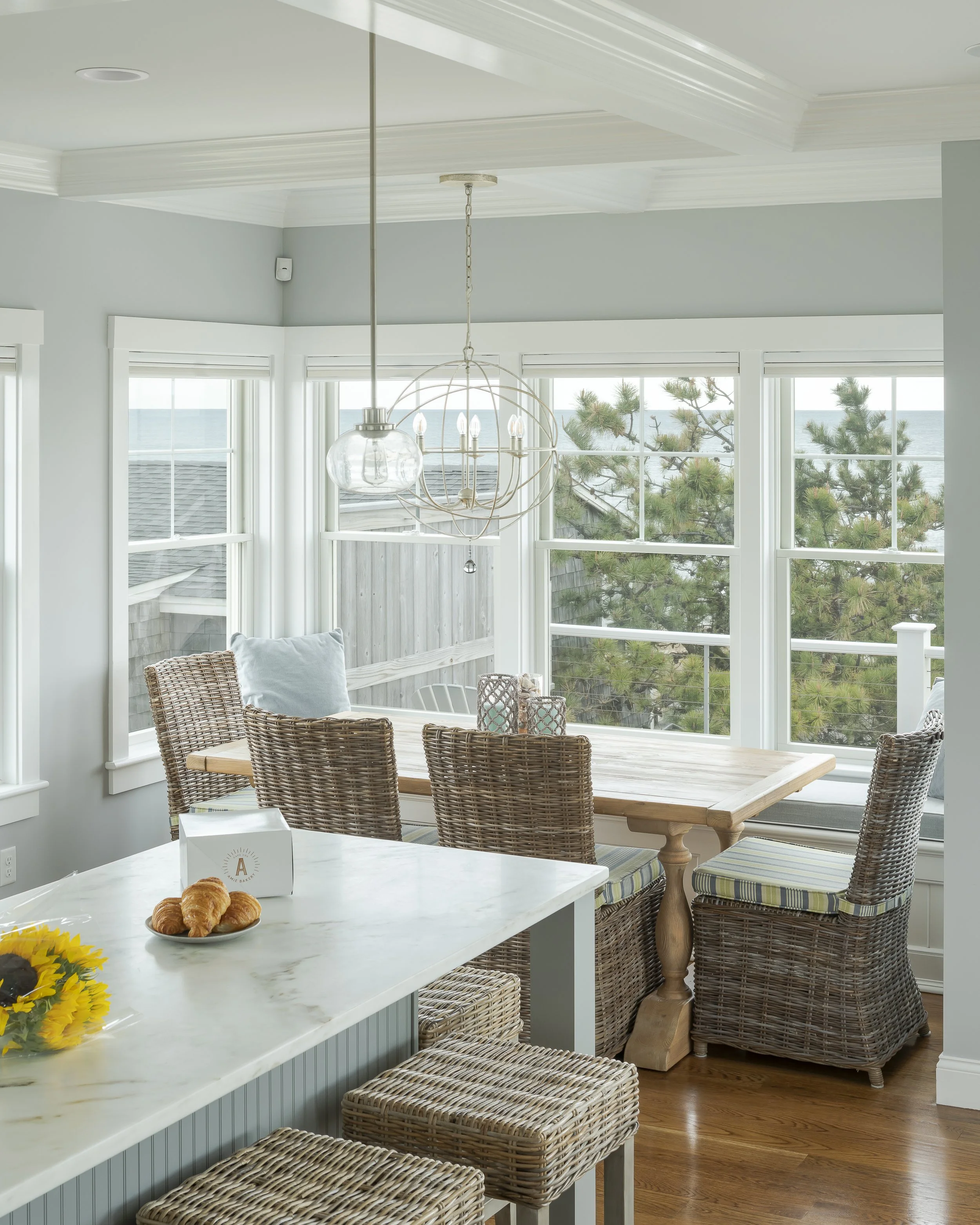 Bright coastal kitchen with a white marble island, wicker chairs around a natural wood table, large windows showing trees and ocean, hanging light fixture, croissants on a plate, and a sunflower in the foreground.