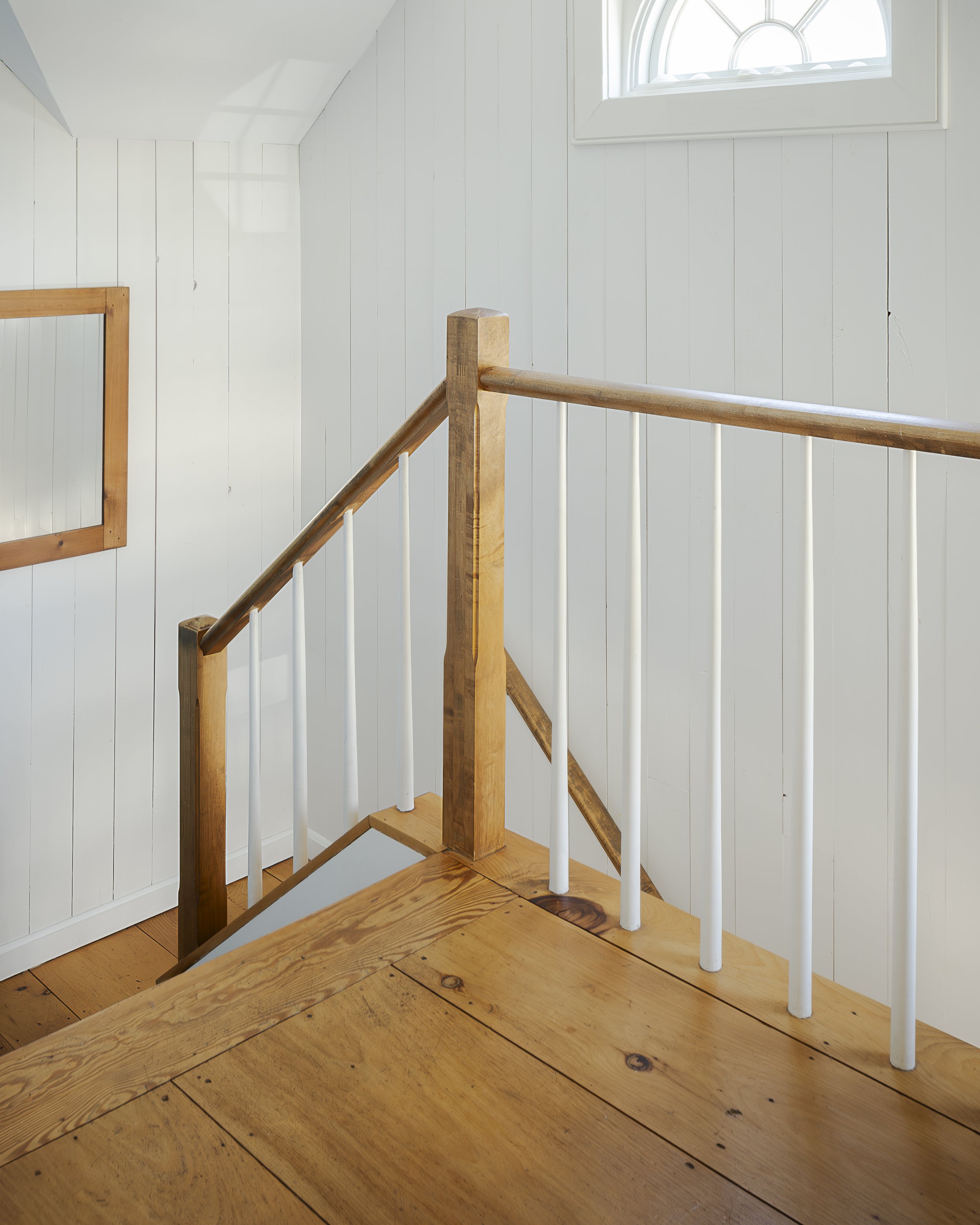 Interior of a white-walled room with a wooden staircase, handrail, and a window letting in natural light.