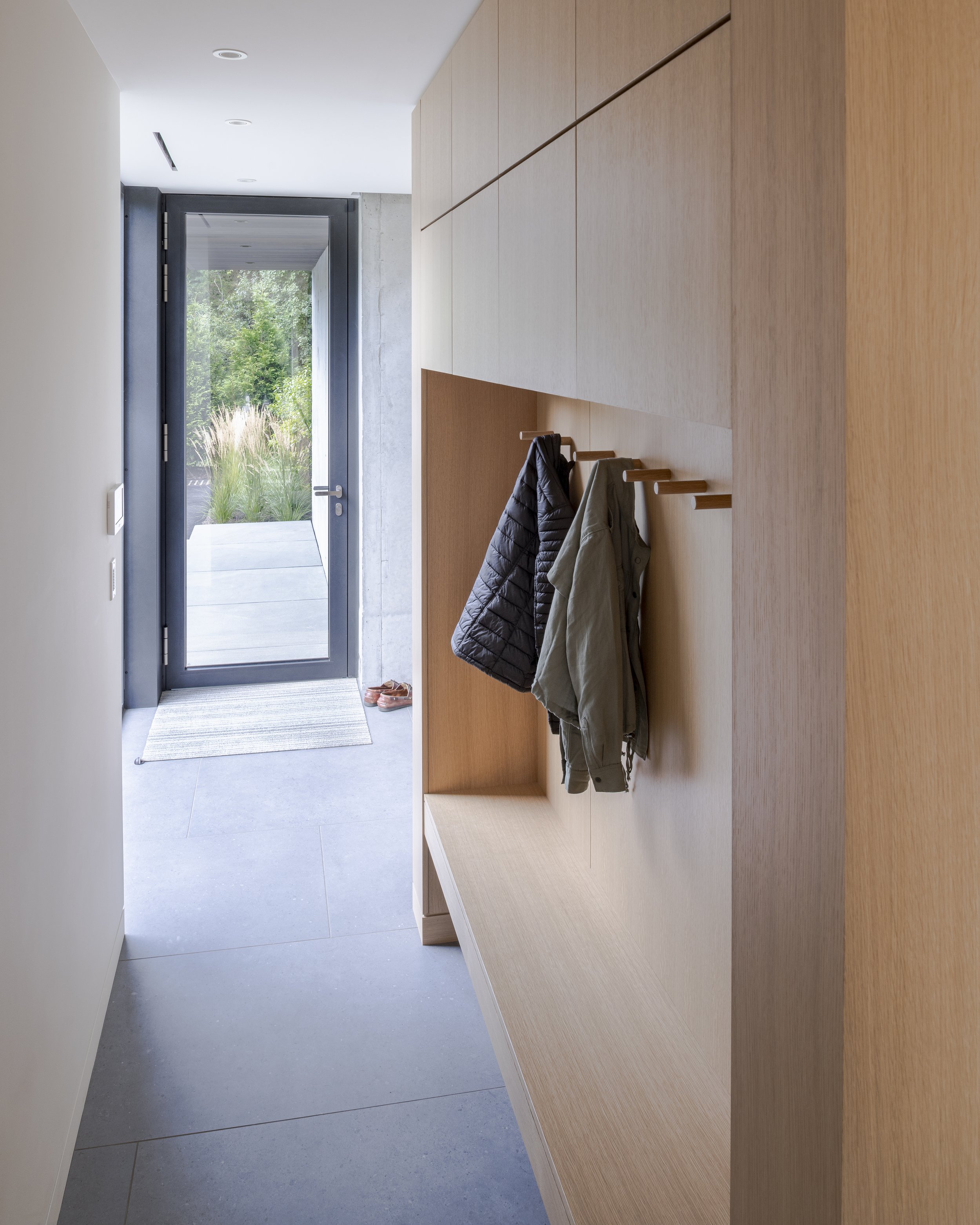Modern entryway with wooden built-in storage and hooks for coats, leading to an outdoor area through a glass door.