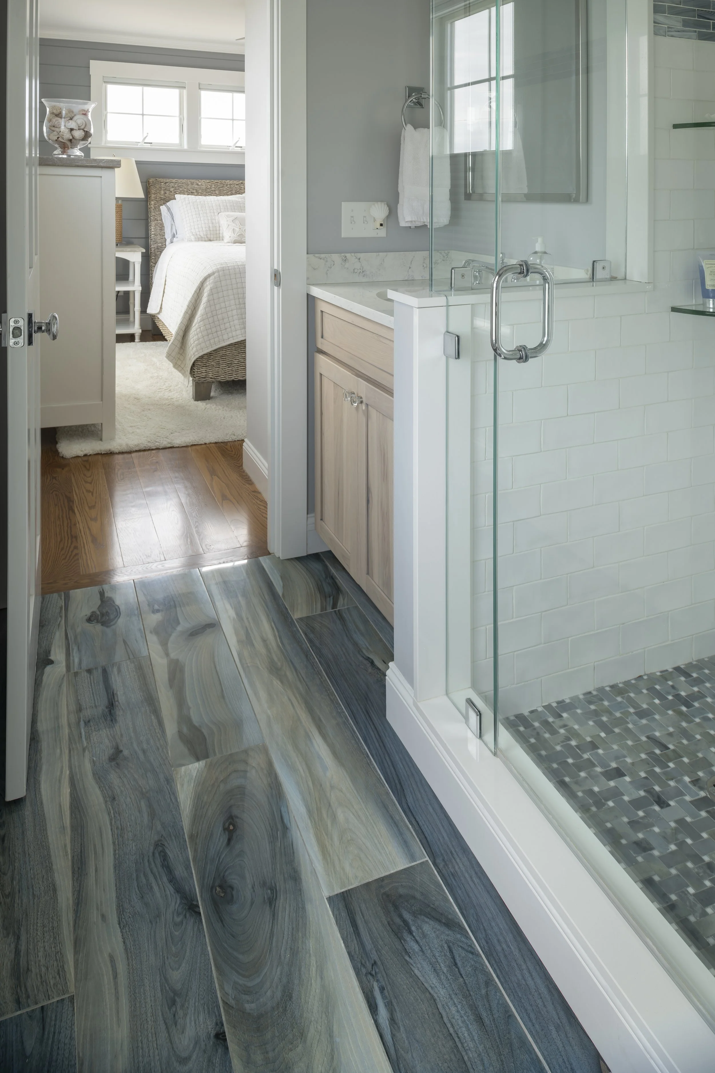 View of a bathroom with a glass shower enclosure, wooden vanity, and a bedroom with a bed and nightstand visible through an open door.