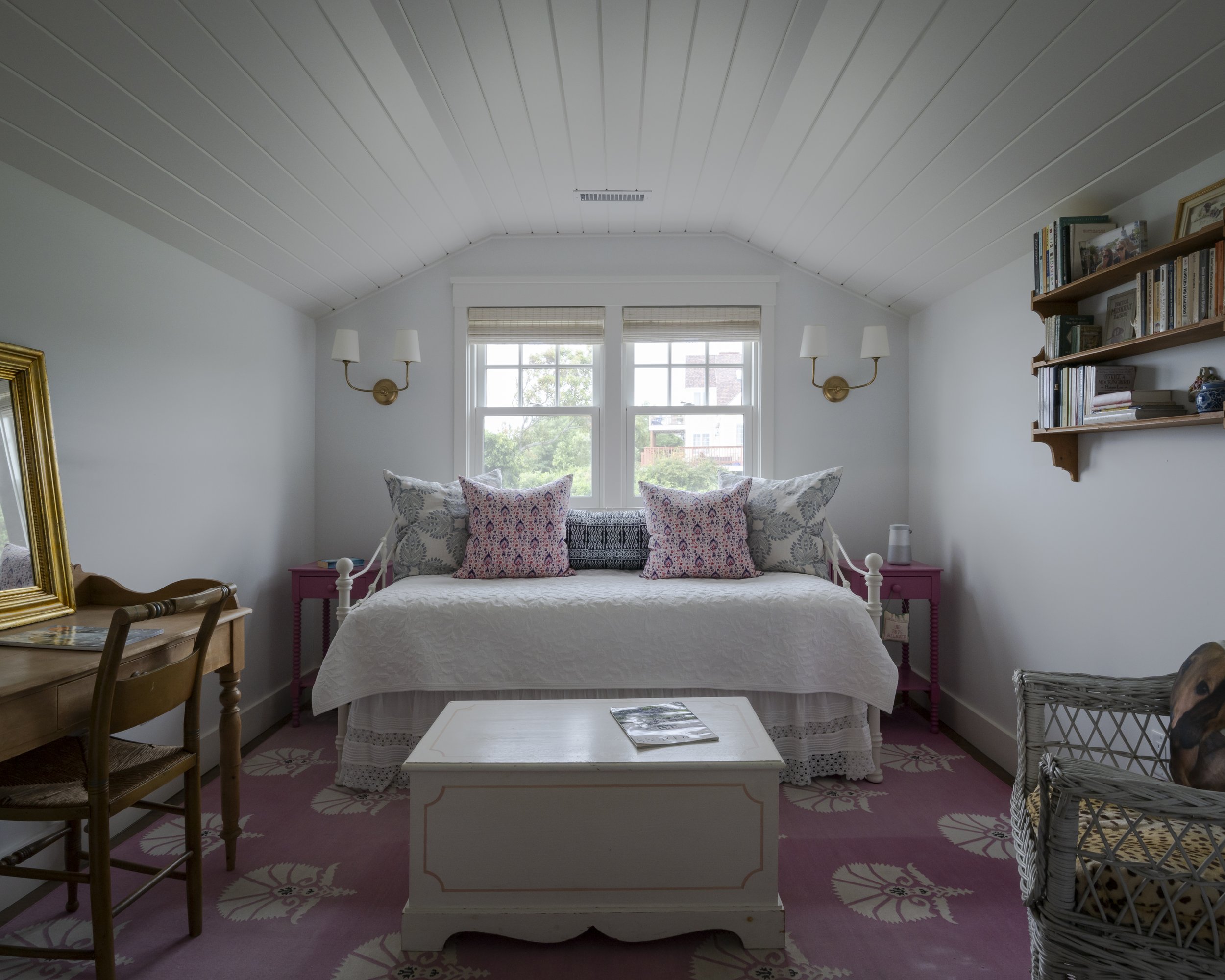 A cozy bedroom with white walls and ceiling, pink floral rug, a white bed with multiple pillows, side tables, a wooden desk with a mirror, a wicker chair, and a bookshelf on the wall.