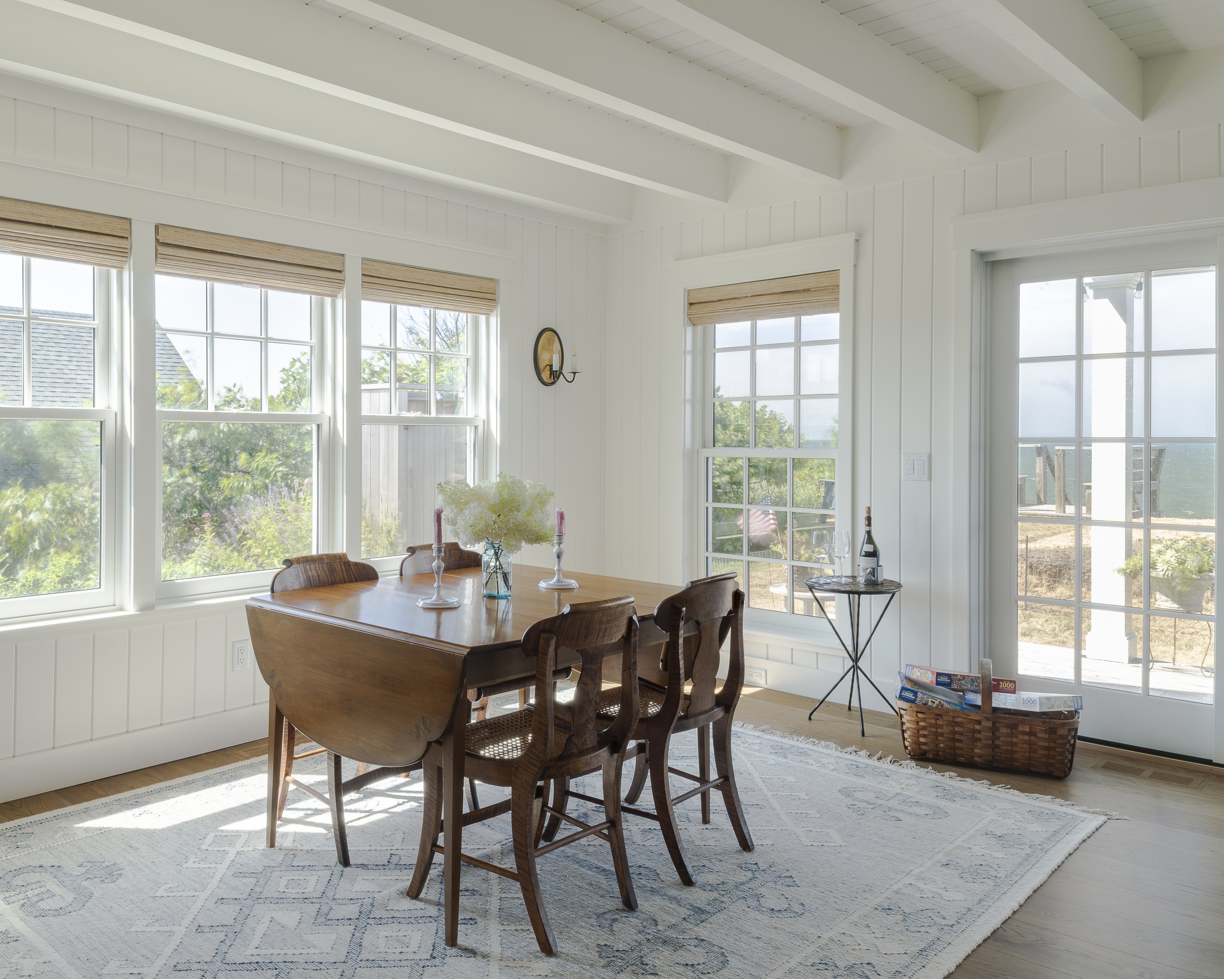 Bright dining room with large windows, white walls, wooden dining table with four chairs, a tray with a wine bottle and glasses, a basket of magazines, and a rug.