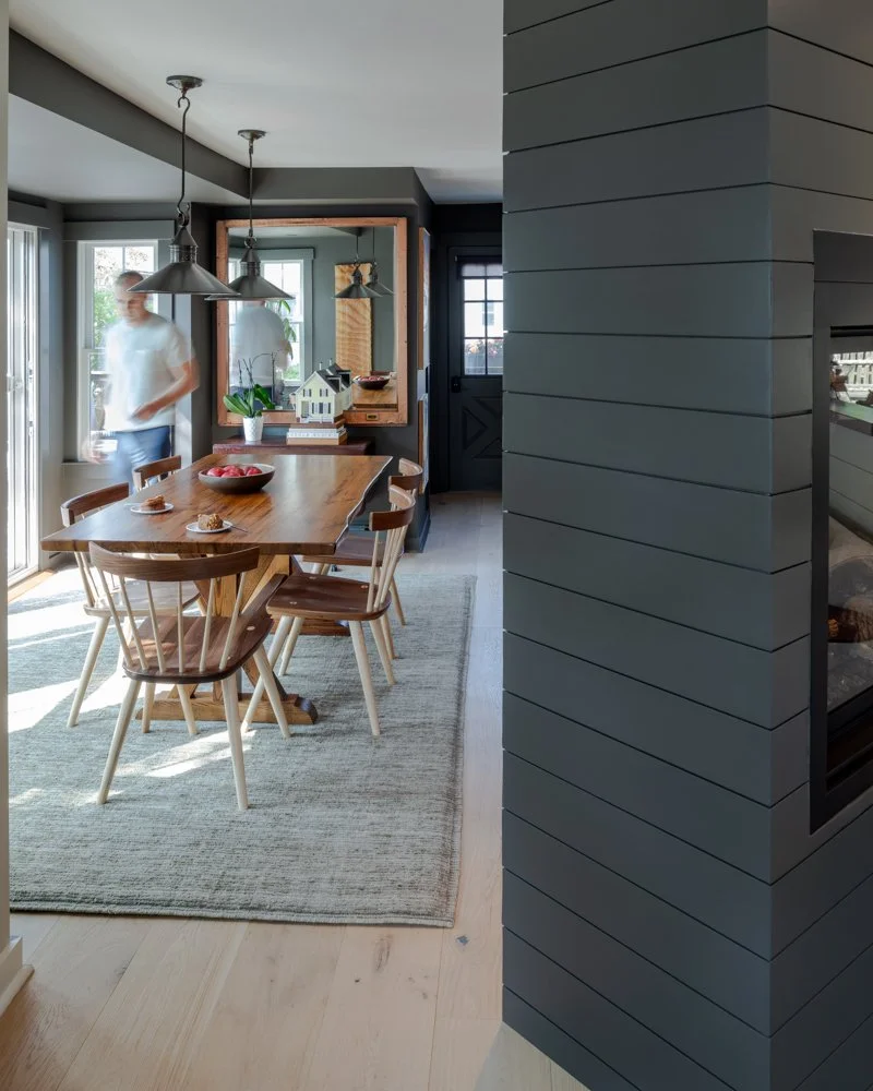 A dining area with a wooden table, six chairs, a bowl of apples, and cookies, illuminated by natural light from sliding glass doors and a window; a person walking past, a mirror on the wall, and a small house model on a side table.