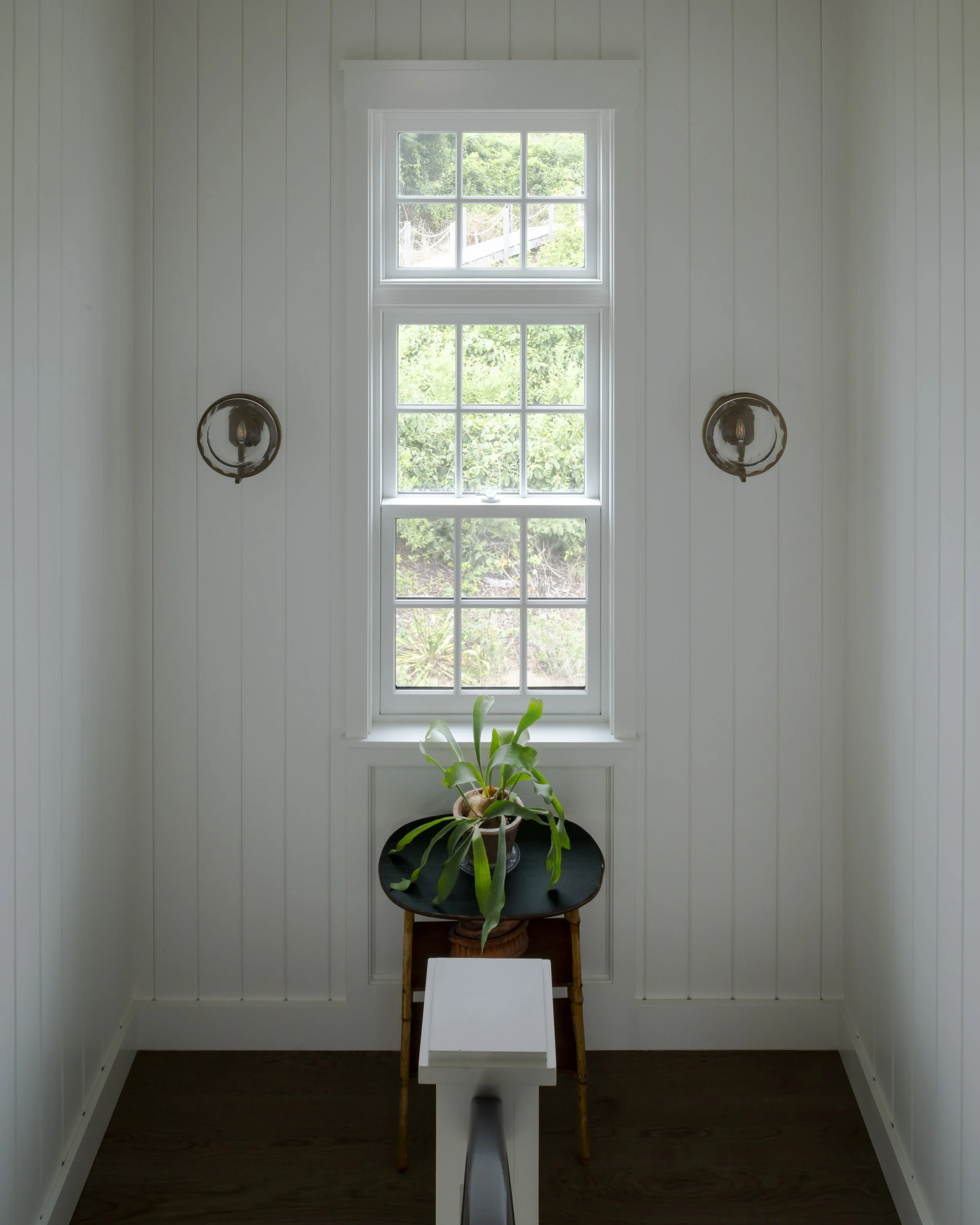 White-walled room with a window, a black table with a potted plant, and wall-mounted lamps.