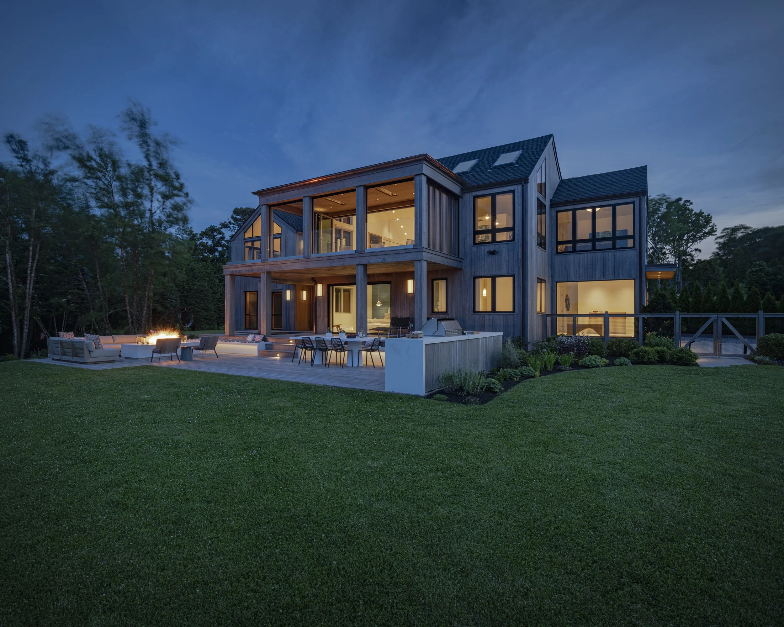 A modern, multi-story house illuminated at dusk with large glass windows, a spacious outdoor patio with seating, a fire pit, and a well-manicured lawn surrounded by trees.