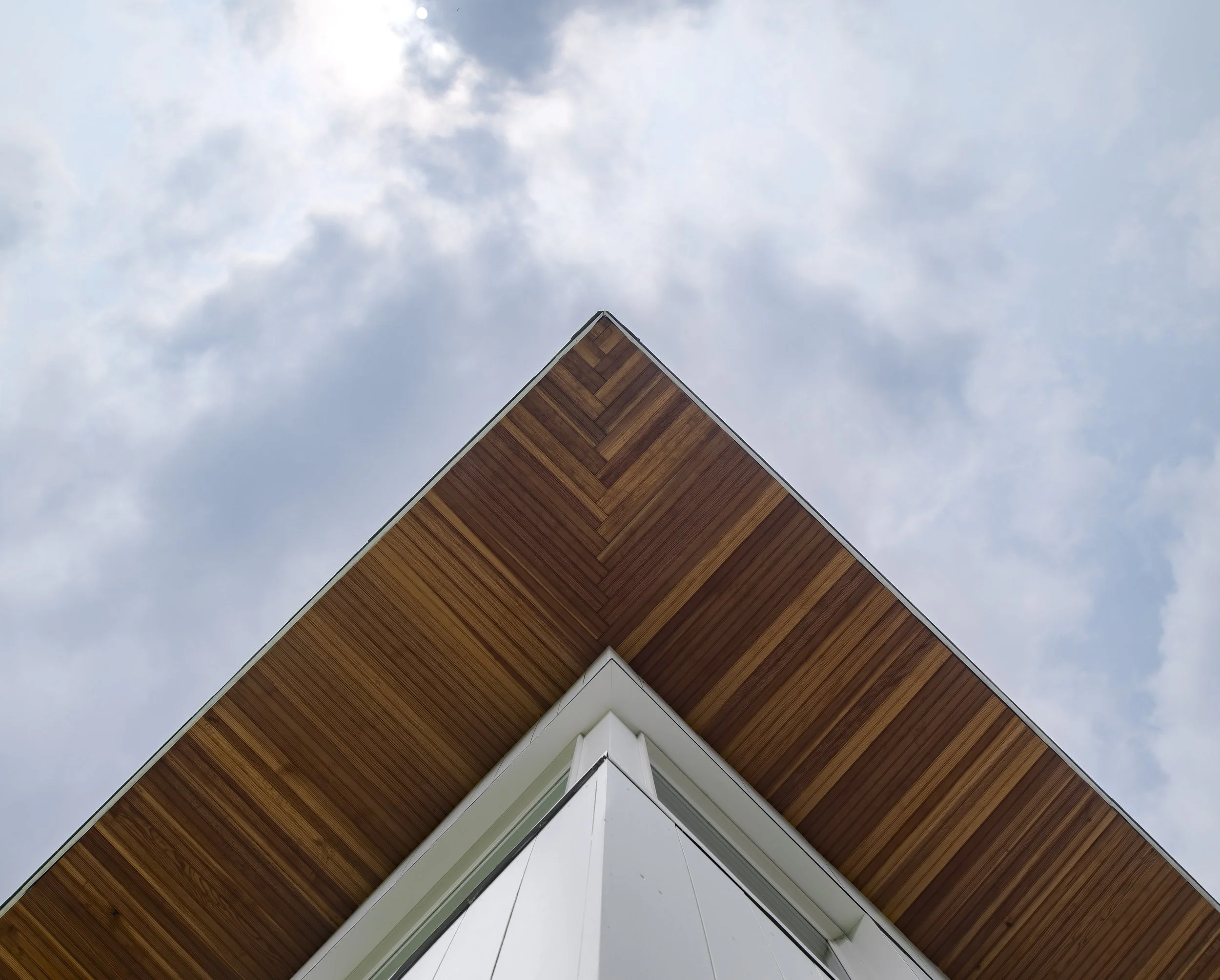 View of the corner of a modern building with a white exterior and a wooden overhang, against a cloudy sky.
