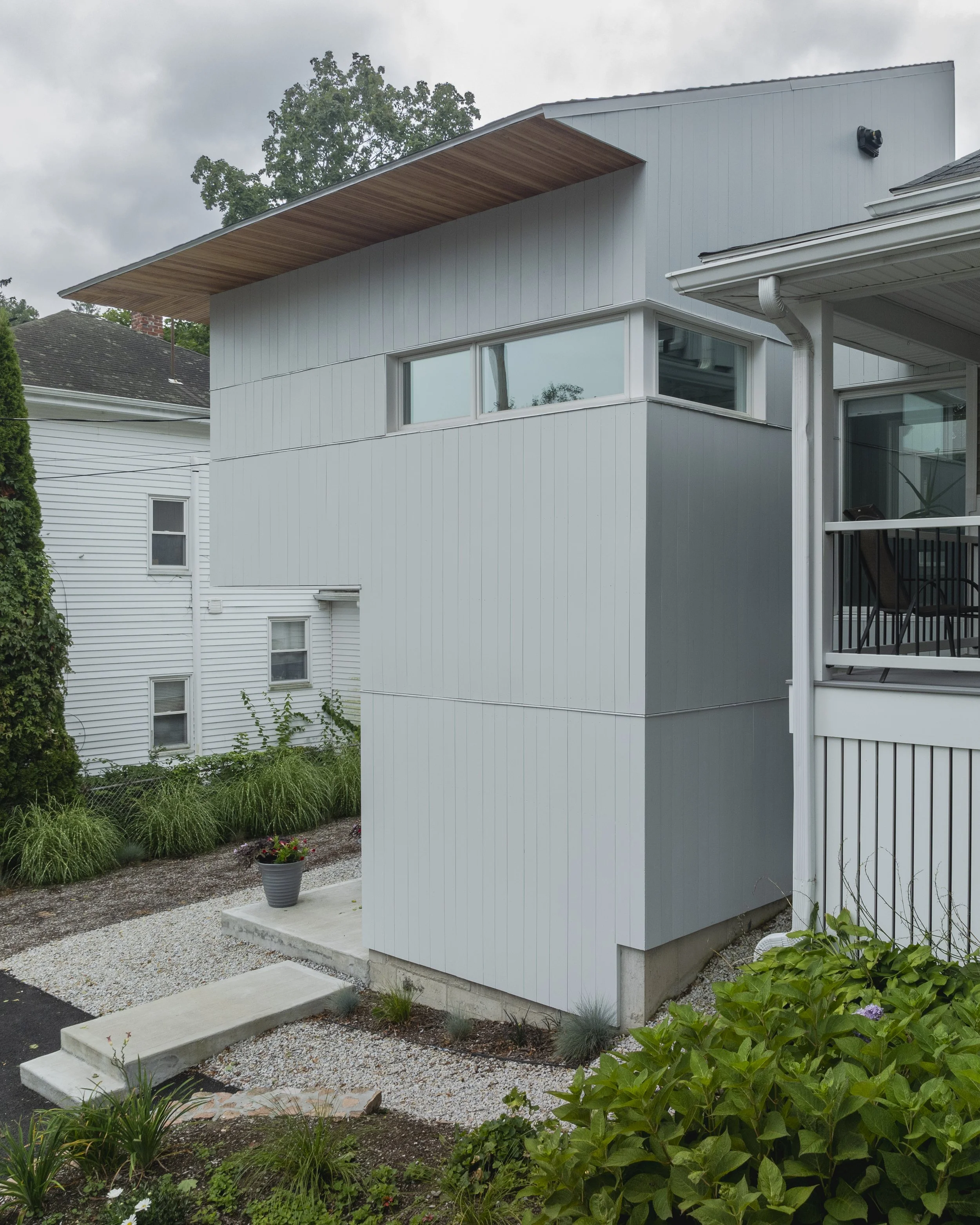Modern white house exterior with large horizontal windows and a wooden overhang, surrounded by a landscaped yard with green plants and a flower pot.