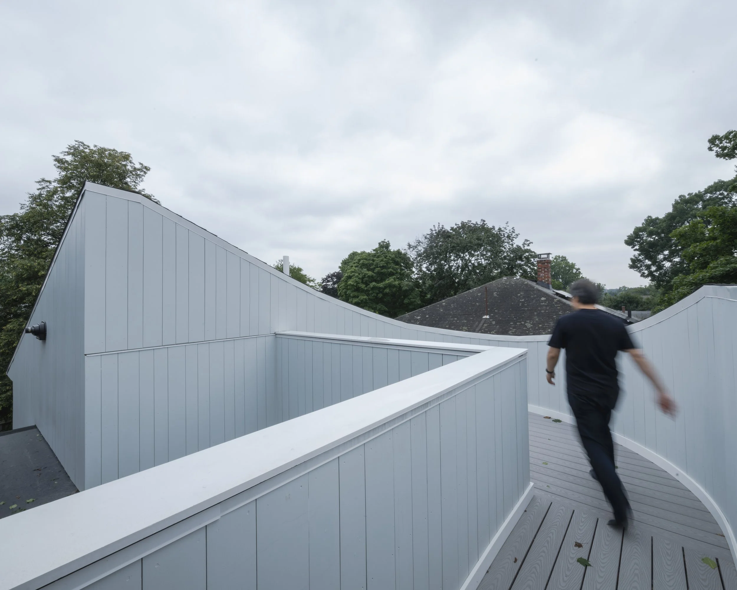 A person walking on a curved, modern rooftop with white siding, surrounded by trees and cloudy sky.