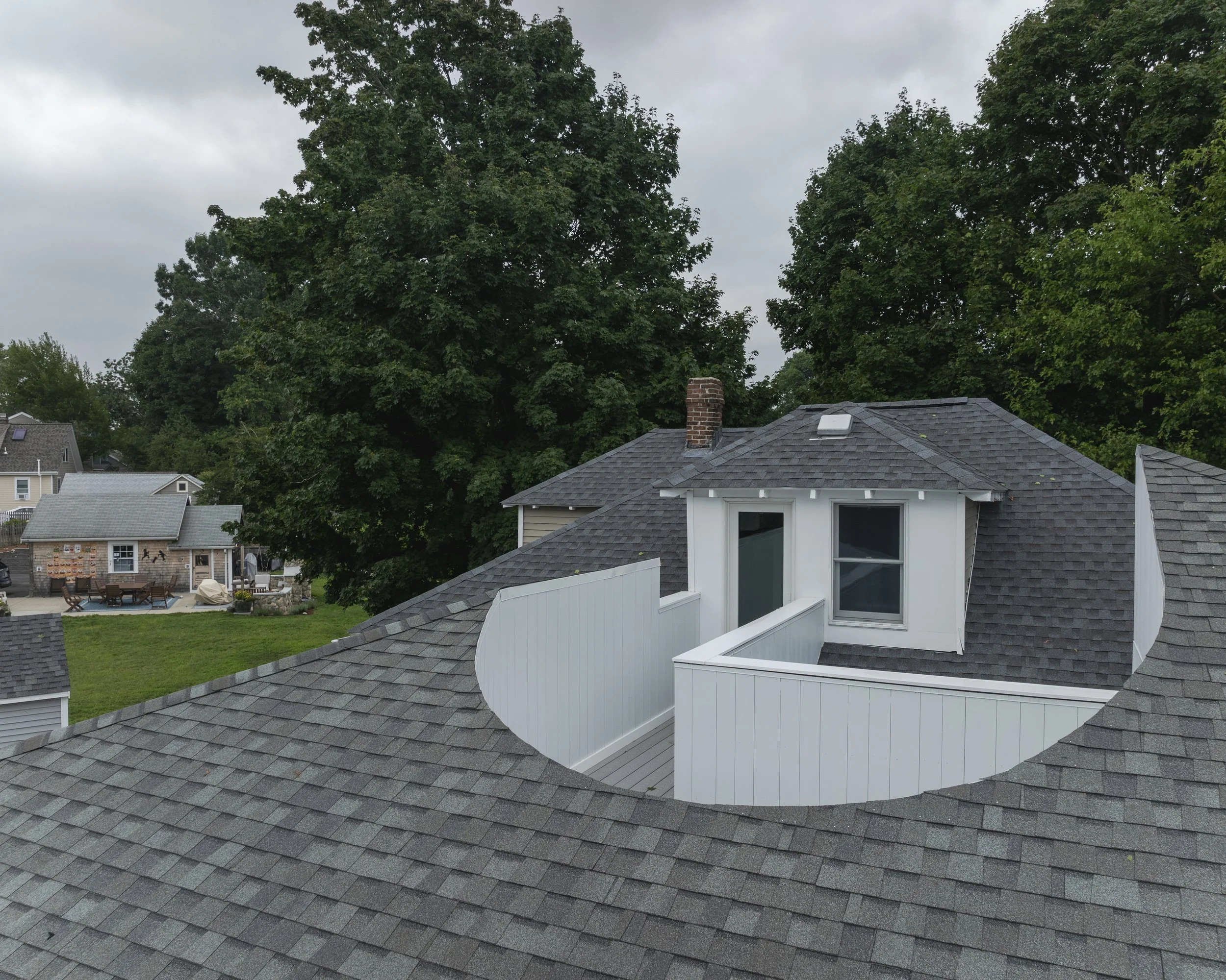A view of a house roof with a small balcony and two windows, surrounded by large green trees and neighboring houses in the background on an overcast day.