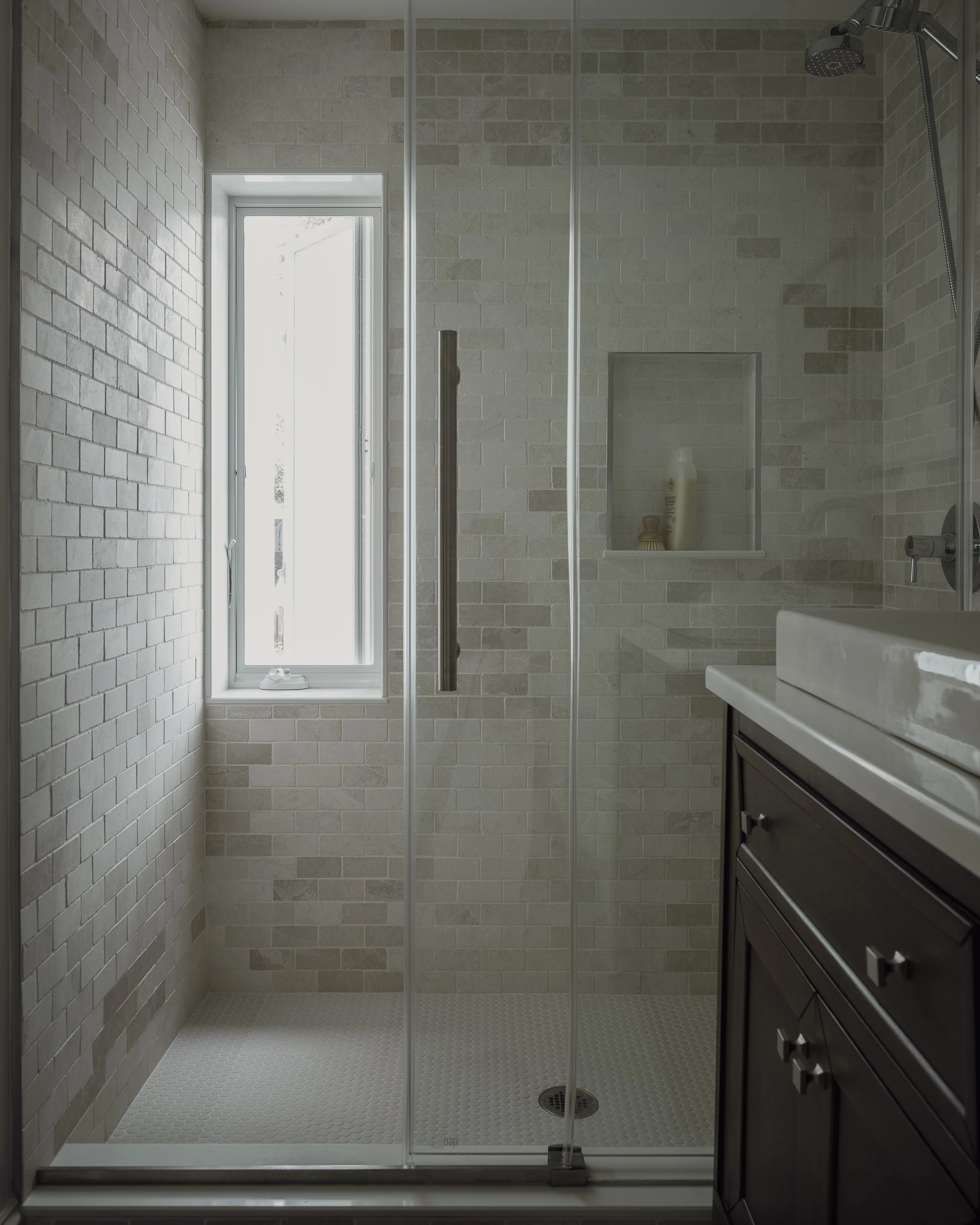 A modern bathroom with a glass-enclosed shower, brick-patterned beige tiles, a narrow window, and a dark wood vanity.