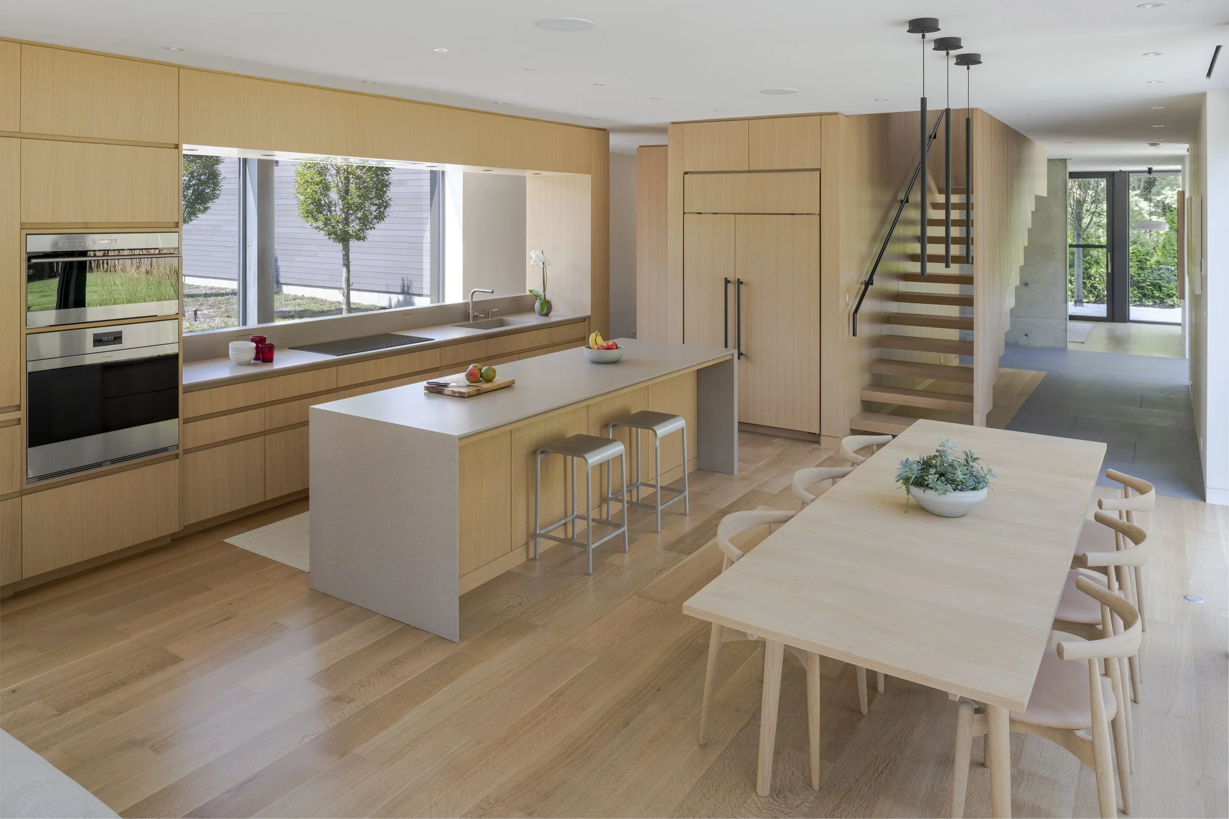 Modern kitchen with light wood cabinetry, a large island with a white countertop, and a dining table with eight chairs. Large window with outdoor views, staircase, and front entrance in the background.