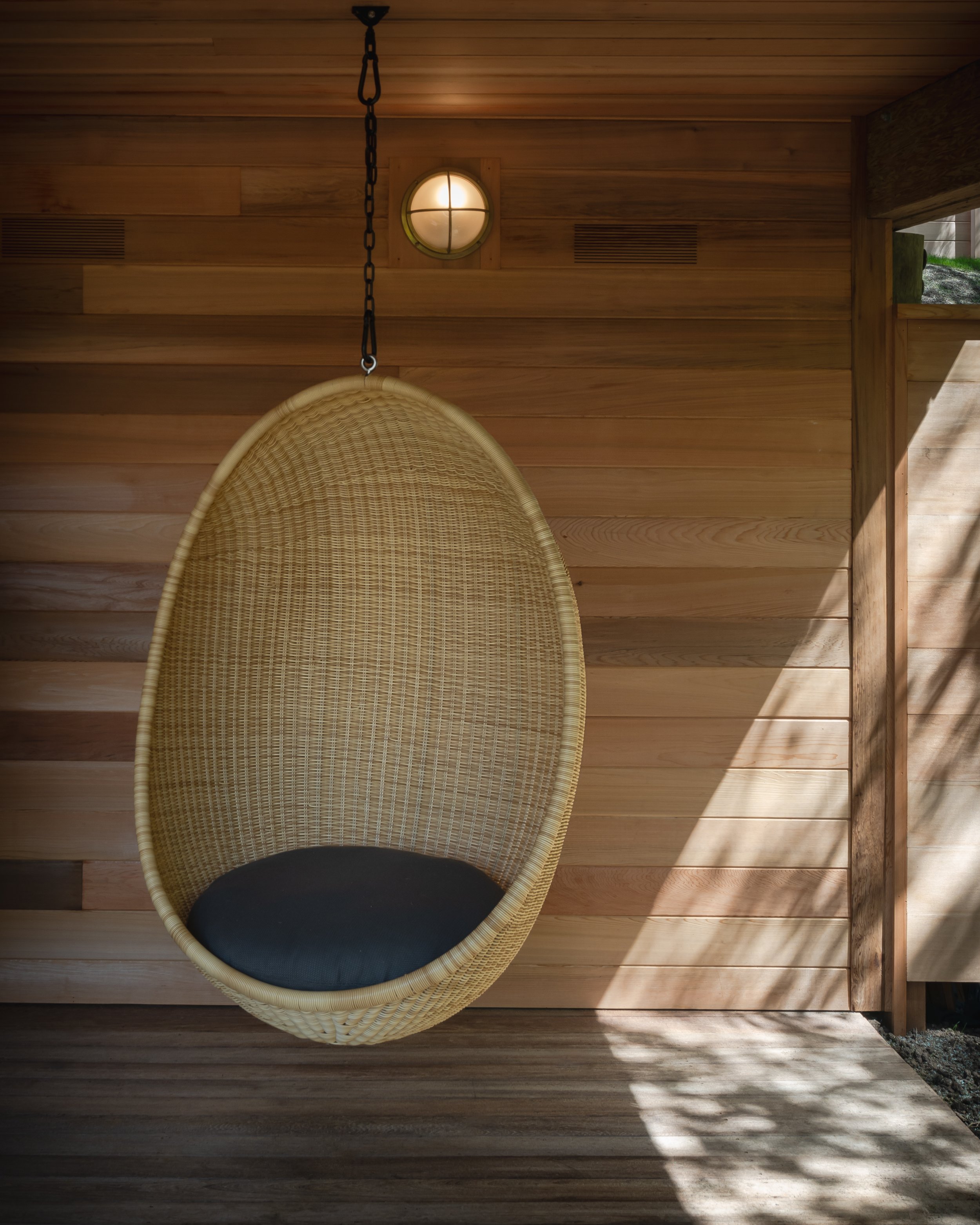 A woven hanging chair with a black cushion, suspended from a chain on a wooden interior wall, with sunlight casting shadows.