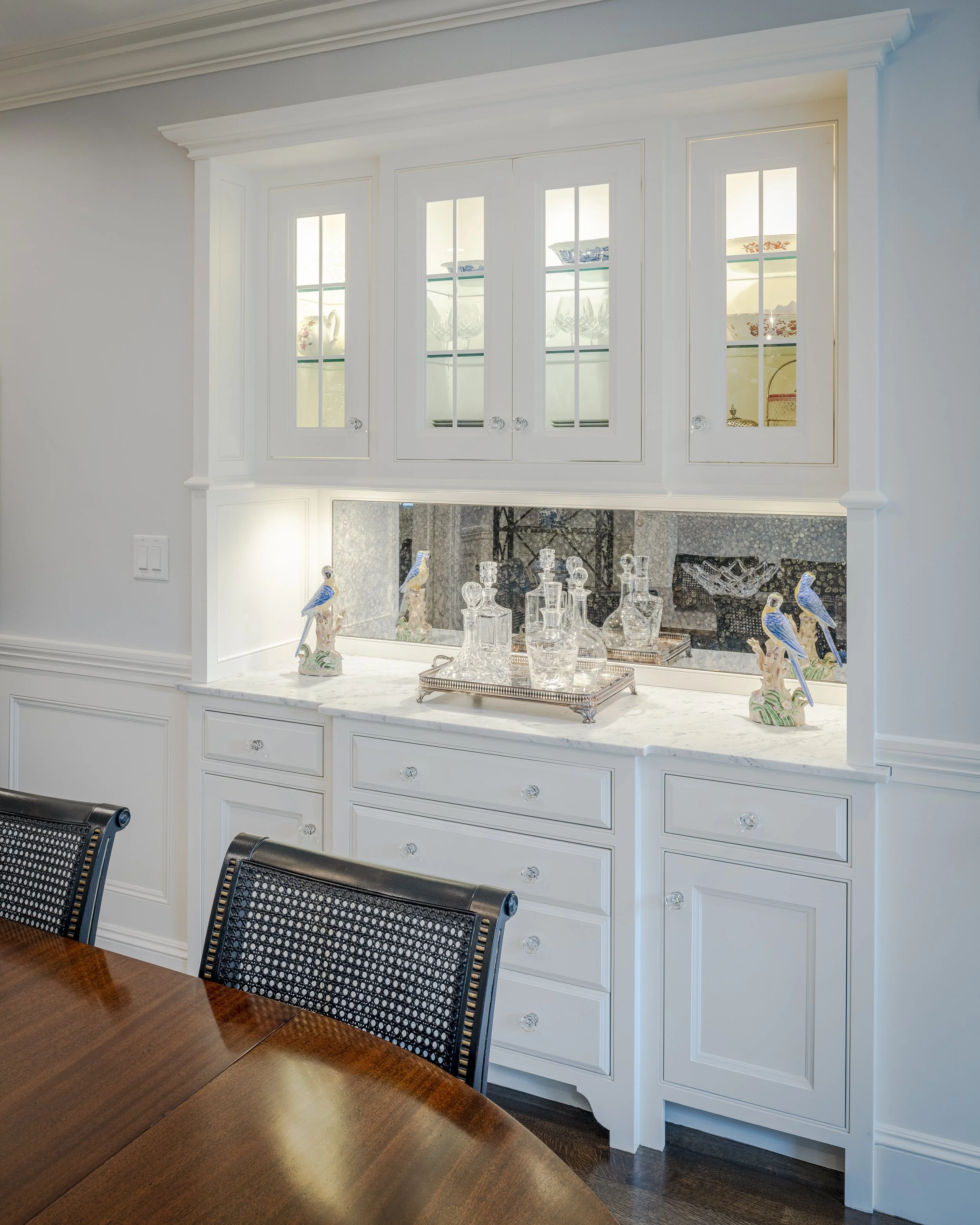 A white built-in china cabinet with glass-front upper cabinets displaying dishes, and a lower section with drawers and cabinets, decorated with glass decanters and porcelain bird figurines, in a dining room.