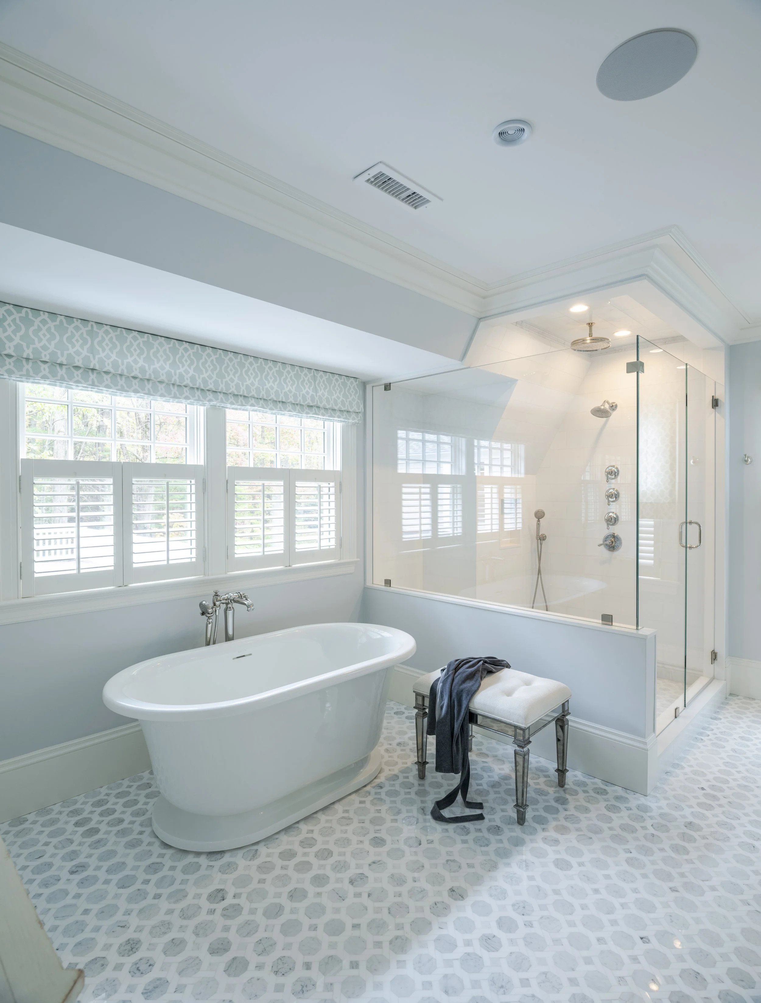 Bright bathroom with white freestanding bathtub, window with patterned valance, and glass-enclosed shower with multiple showerheads, decorated in light blue and white tones.