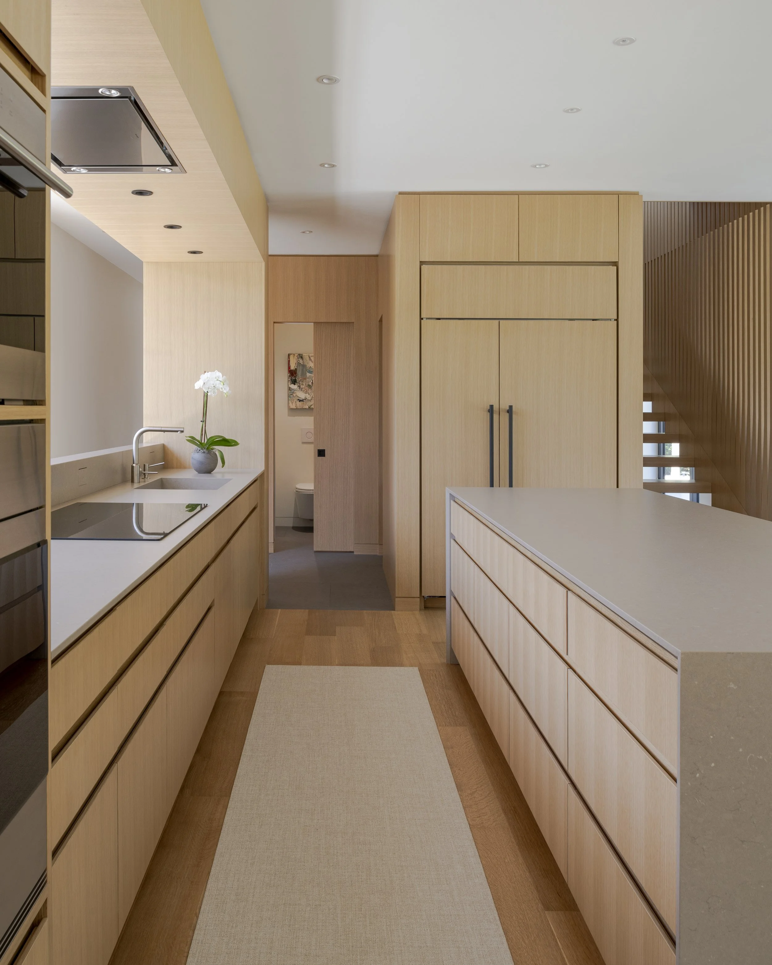 Modern kitchen with light wood cabinets, a white countertop, a built-in stovetop, a potted plant, and a view into a bathroom with a toilet.