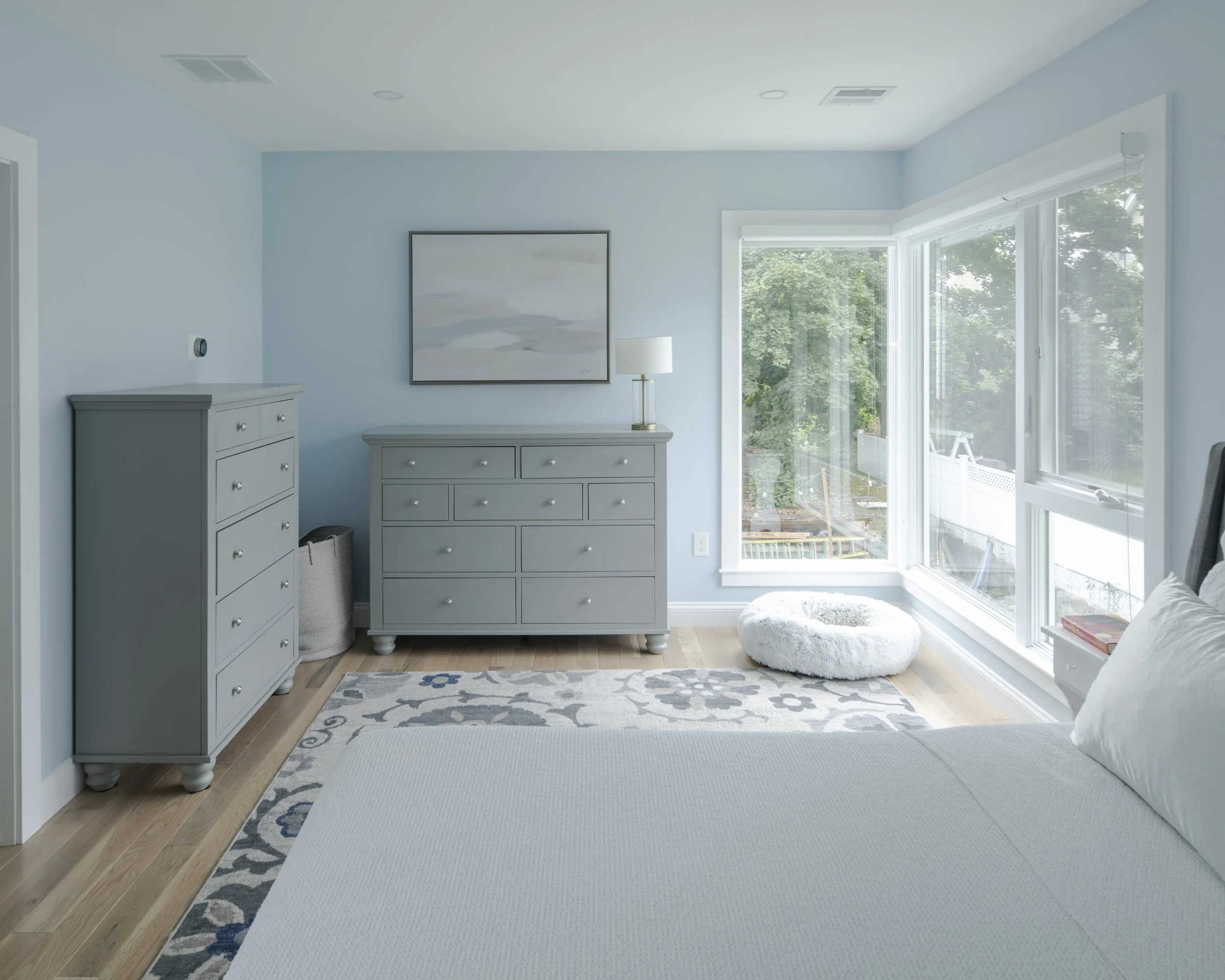 A bright bedroom with light blue walls, white bedding, a large window with a view of trees, a gray dresser, a matching gray chest of drawers, a white fluffy pet bed near the window, a table lamp on the dresser, a framed abstract painting, and a patte