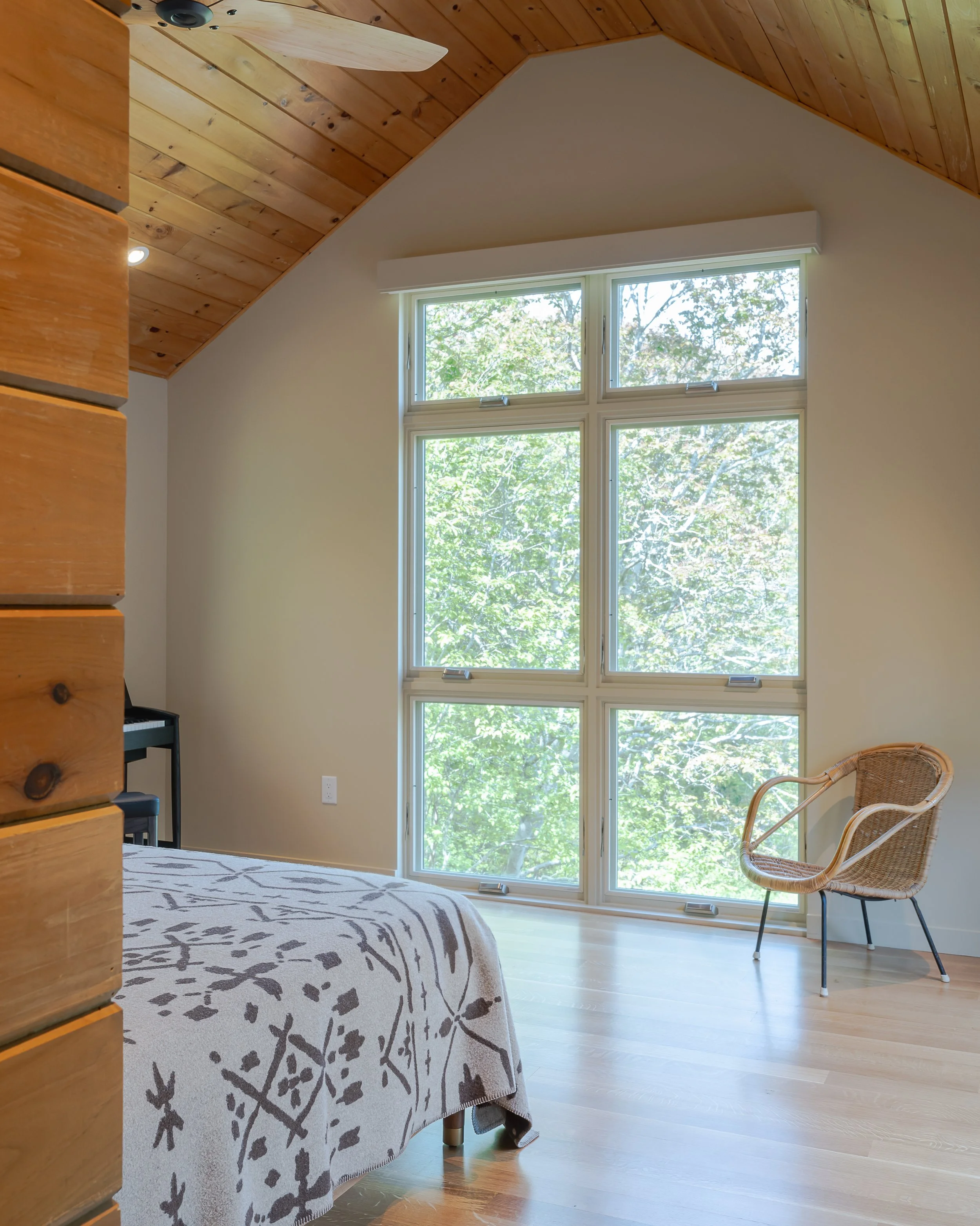 A bedroom with a large window showing green trees outside, a bed with a patterned blanket, a wicker chair, a wooden wall, and a ceiling with wood paneling.