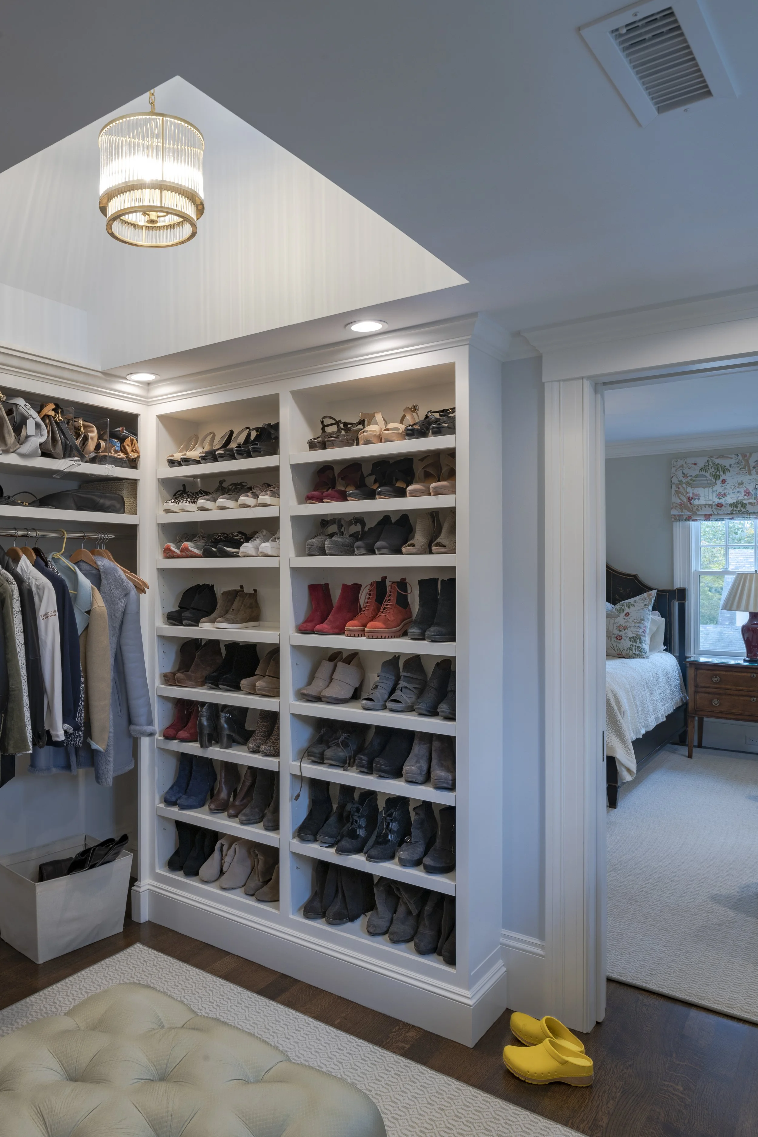 A walk-in closet with white built-in shelving filled with various shoes and boots, a clothing rack with jackets and shirts, a beige ottoman at the bottom, and a pair of yellow slippers on the dark hardwood floor, with a bedroom visible through an ope