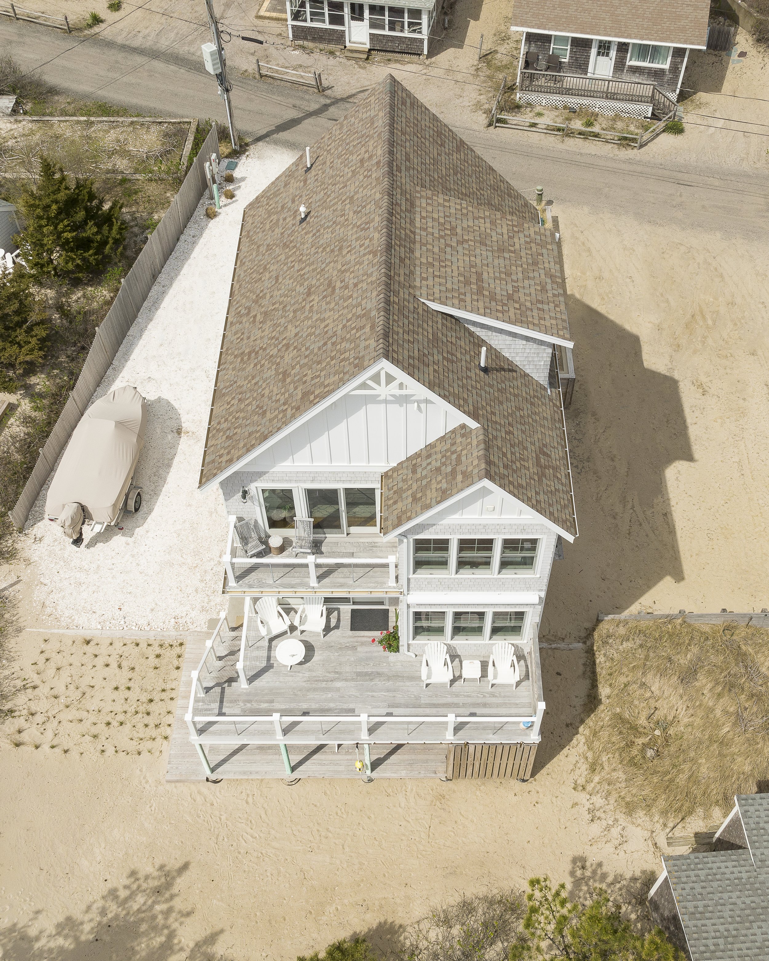 An aerial view of a house with a brown shingle roof, white exterior walls, and a large wooden deck with white patio chairs and a round table. The yard is mostly sandy with some grass and a small garden bed.