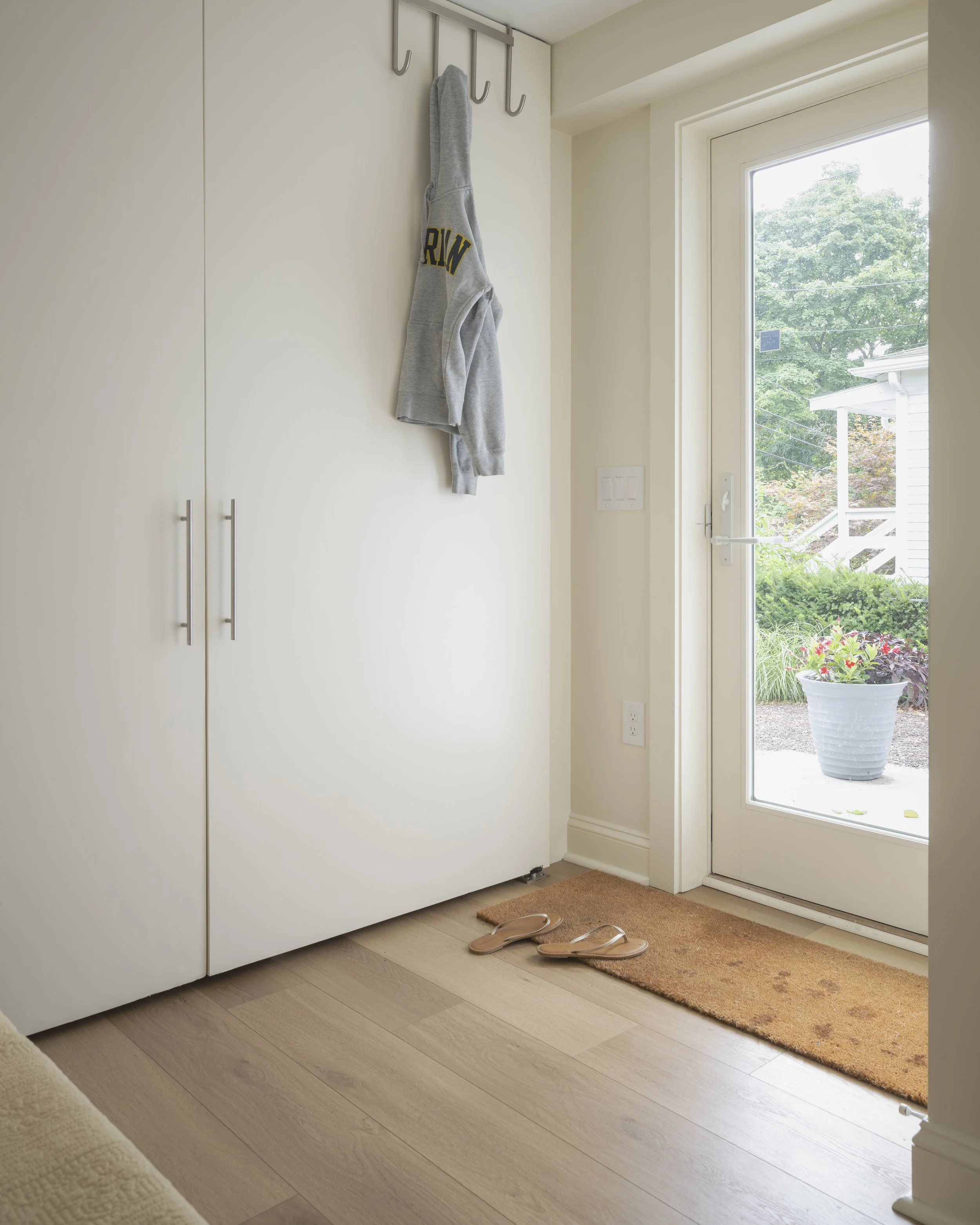 Entryway with a white cabinet, a gray sweatshirt hanging on hooks, a brown doormat, and pink sandals near the glass door with a view of a potted plant outside.