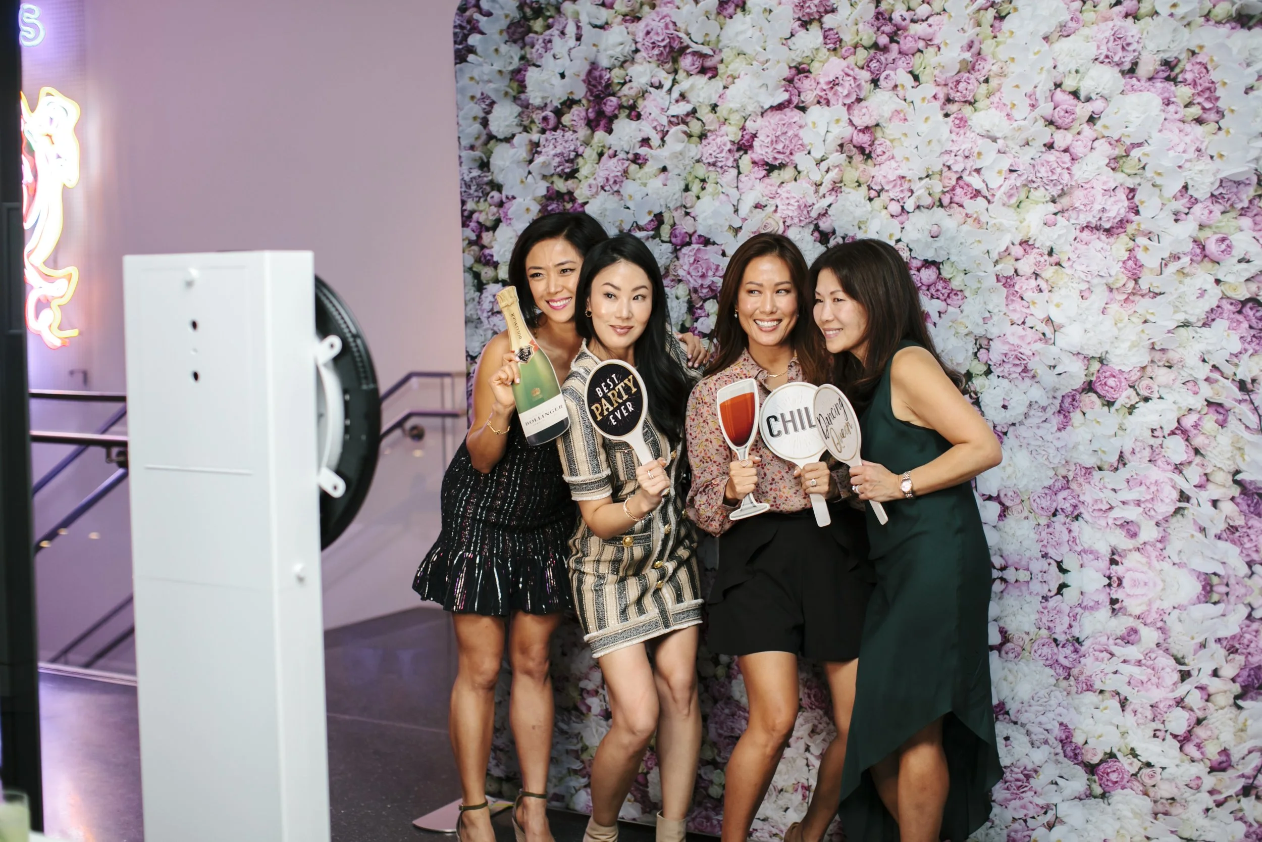 Four women are smiling and posing for a photo at a party, holding various drink-themed signs, in front of a floral backdrop.