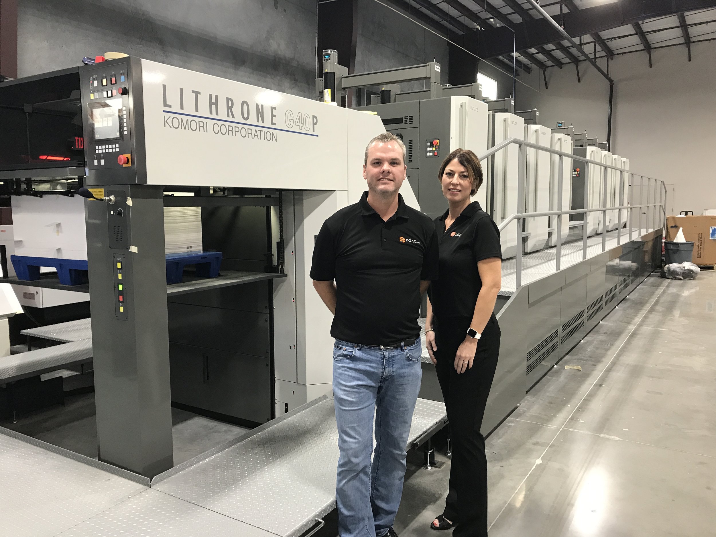 Shown here, Todd and Brandy Frost of Next Day Four Color pause in front of Komori press that is equipped with XO Series UV-LED technology from AMS Spectral UV - A Baldwin Technology Company.