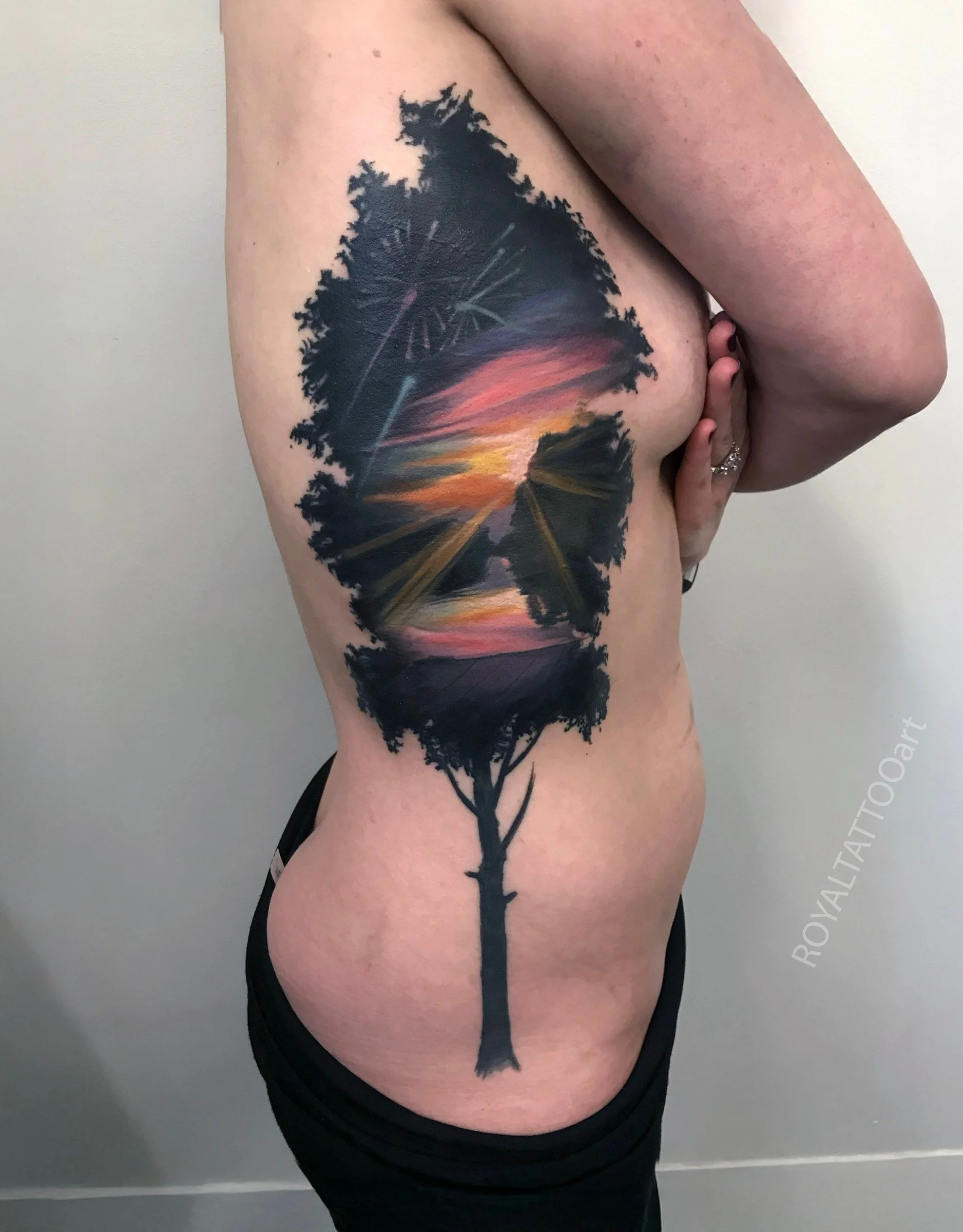 A double-exposure tattoo featuring a tree silhouette within the outline of a leaf, blending nature and sunset colors along the side of the torso.