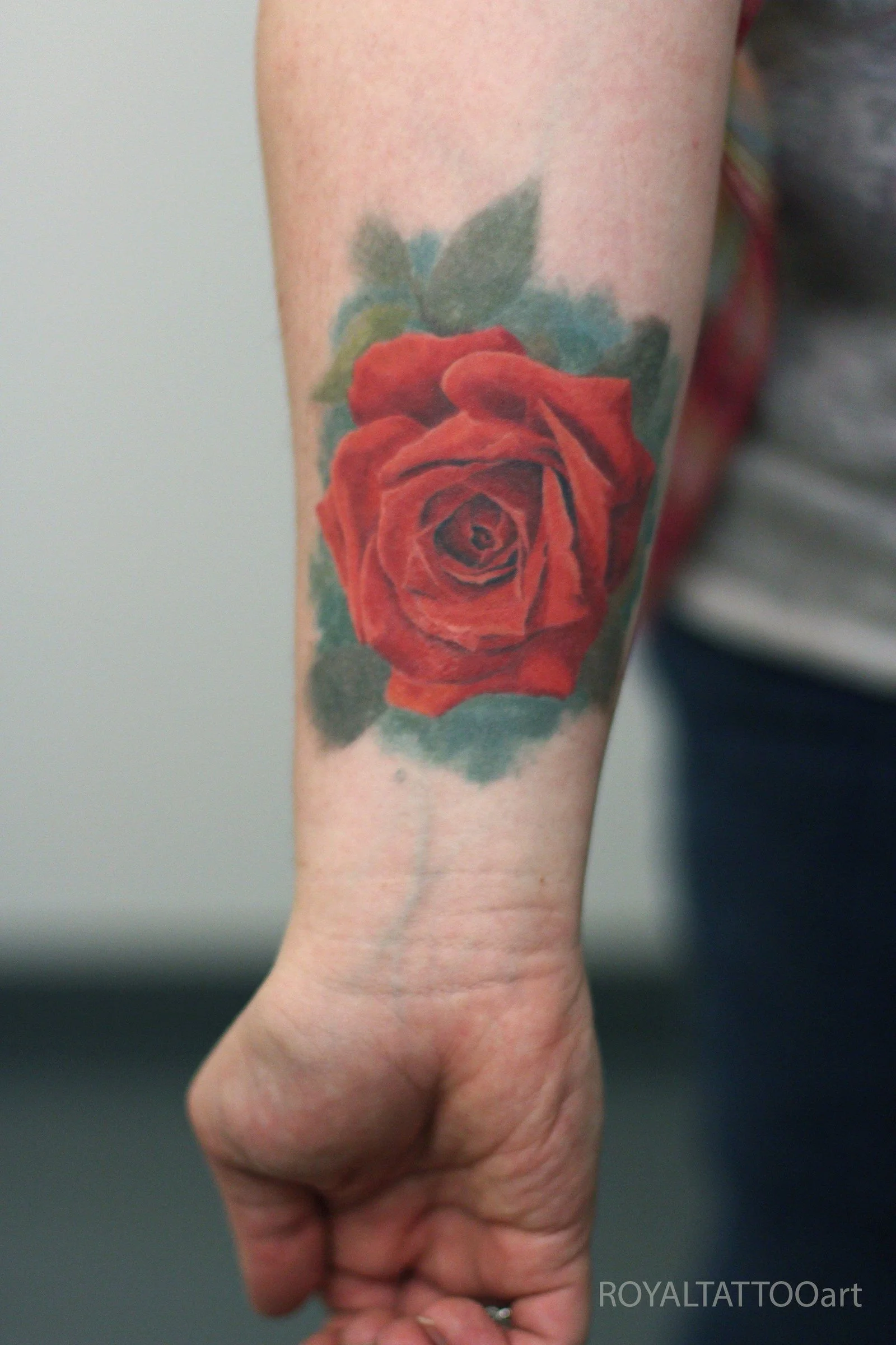 A realistic color rose tattoo with rich red petals and subtle green leaves, placed prominently on the forearm.
