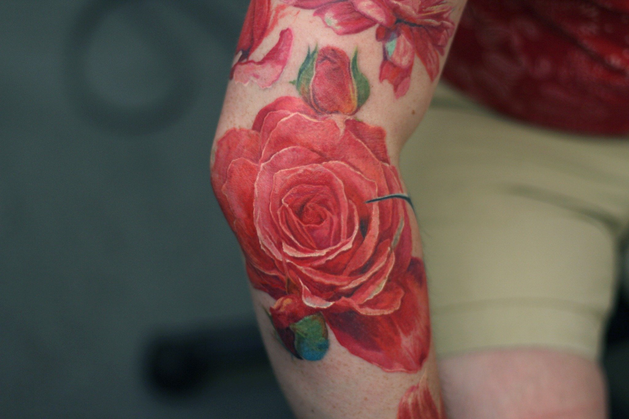 A realistic color rose tattoo on the thigh features a large red bloom with delicate petals and lush green leaves.