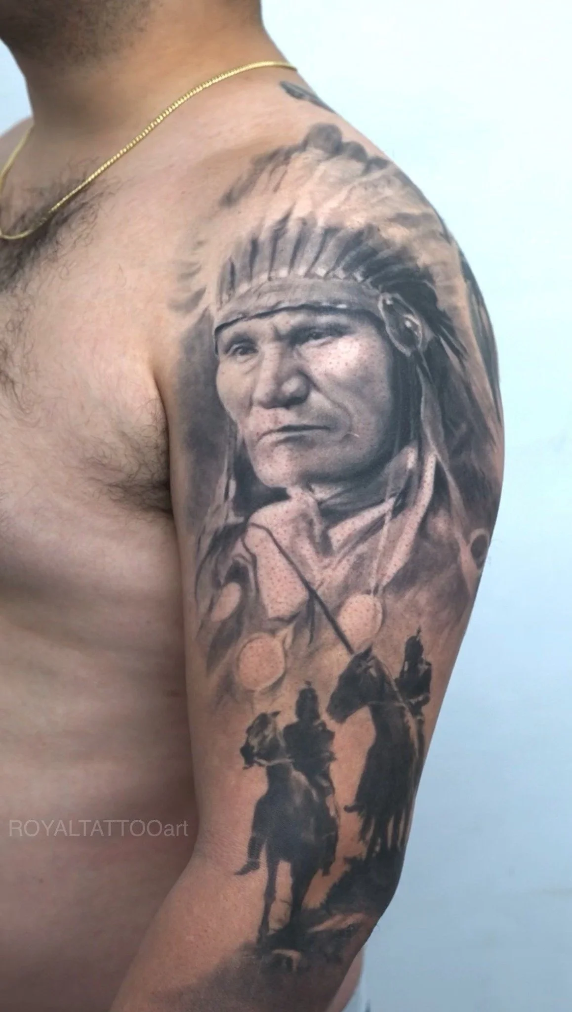 Black and grey Native chief portrait tattoo on the upper arm, featuring horseback rider details and realistic shading by Royal Jafarov in NYC.
