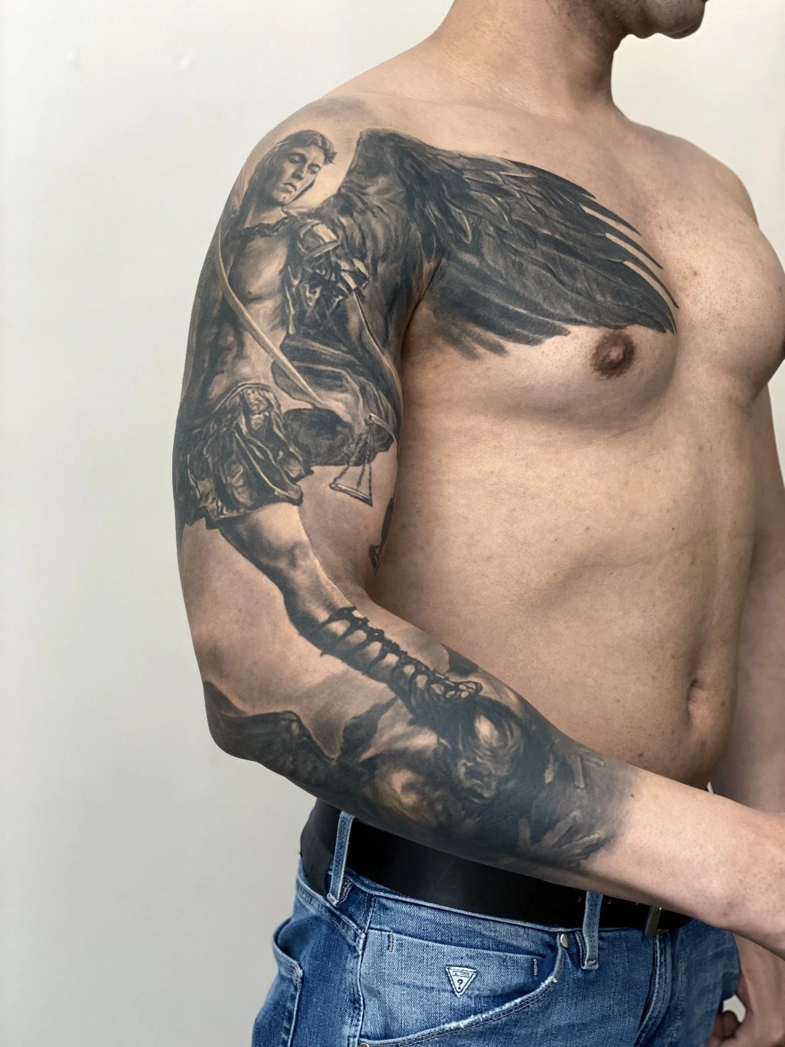 Black and grey religious realism upper arm tattoo featuring an angel warrior with detailed wings, armor, and dramatic shading.
